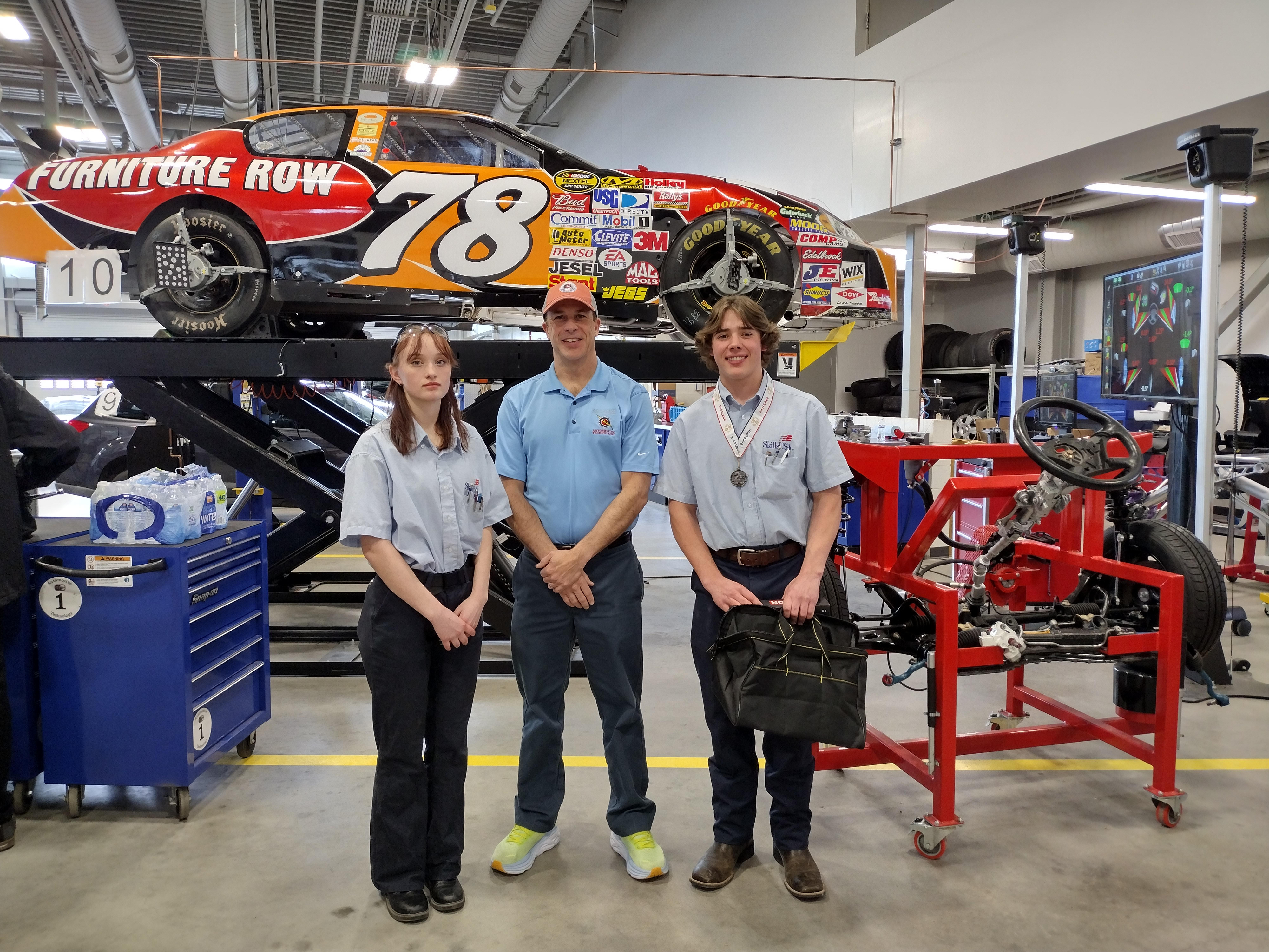 A female student, an instructor, and a male student wearing auto shop clothing stand in front of an elevated car with "Furniture Row" and number 78 on it.
