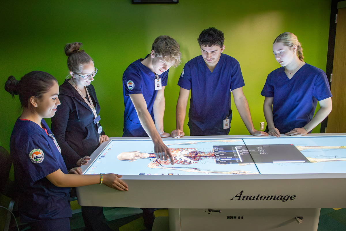 A group of 5 health sciences students, 3 female and 2 male, are wearing blue scrubs and studying a virtual cadaver on EPIC Campus's Anatomage table.