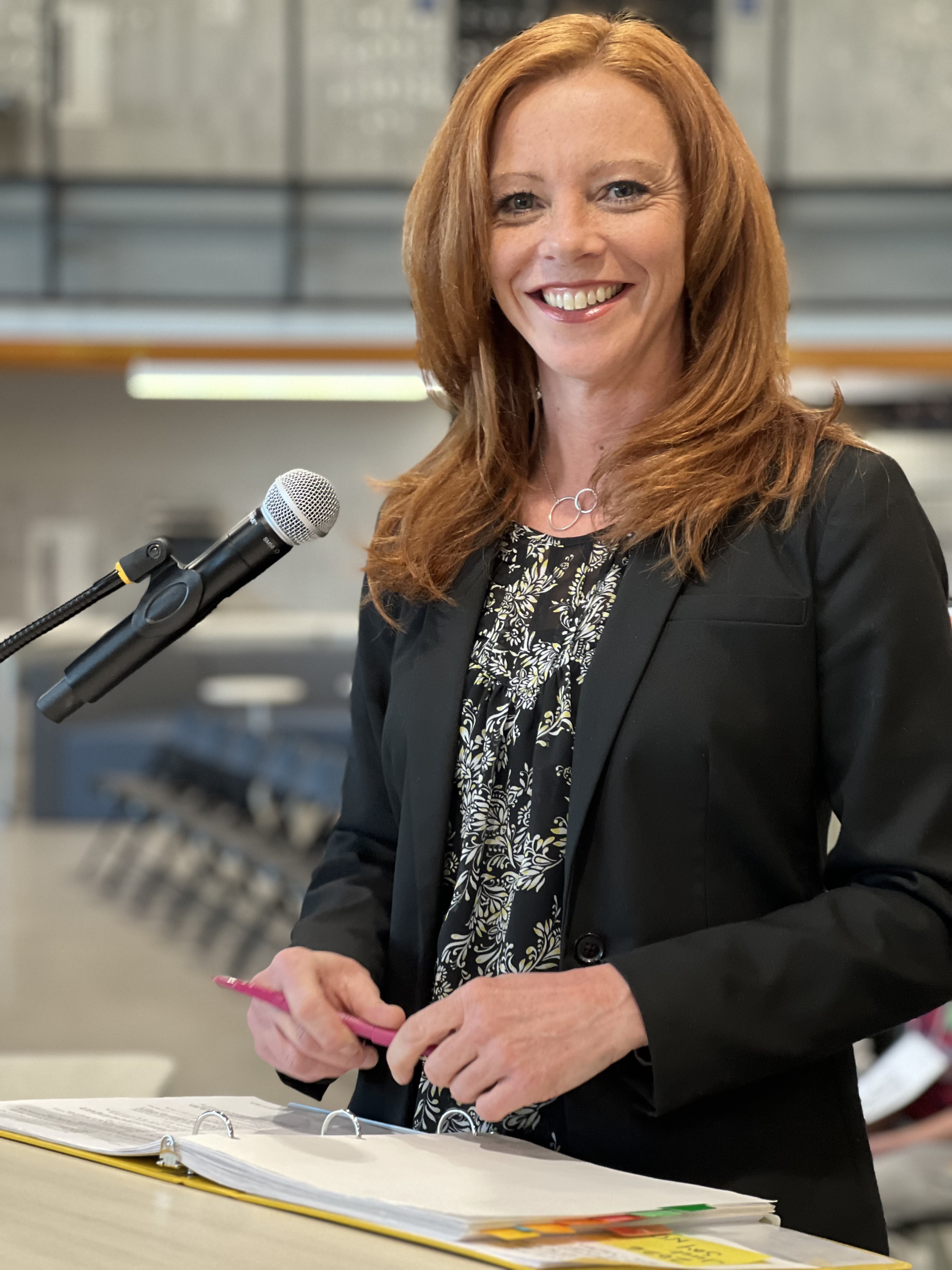 The image shows a woman with shoulder-length red hair and a bright smile standing at a podium. She is wearing a black blazer over a black and white floral blouse and holds a pink pen in her hand. A microphone is positioned in front of her, and an open binder with papers is on the podium. The background features an indoor setting with chairs arranged in rows, suggesting a formal event or competition, likely a spelling bee.