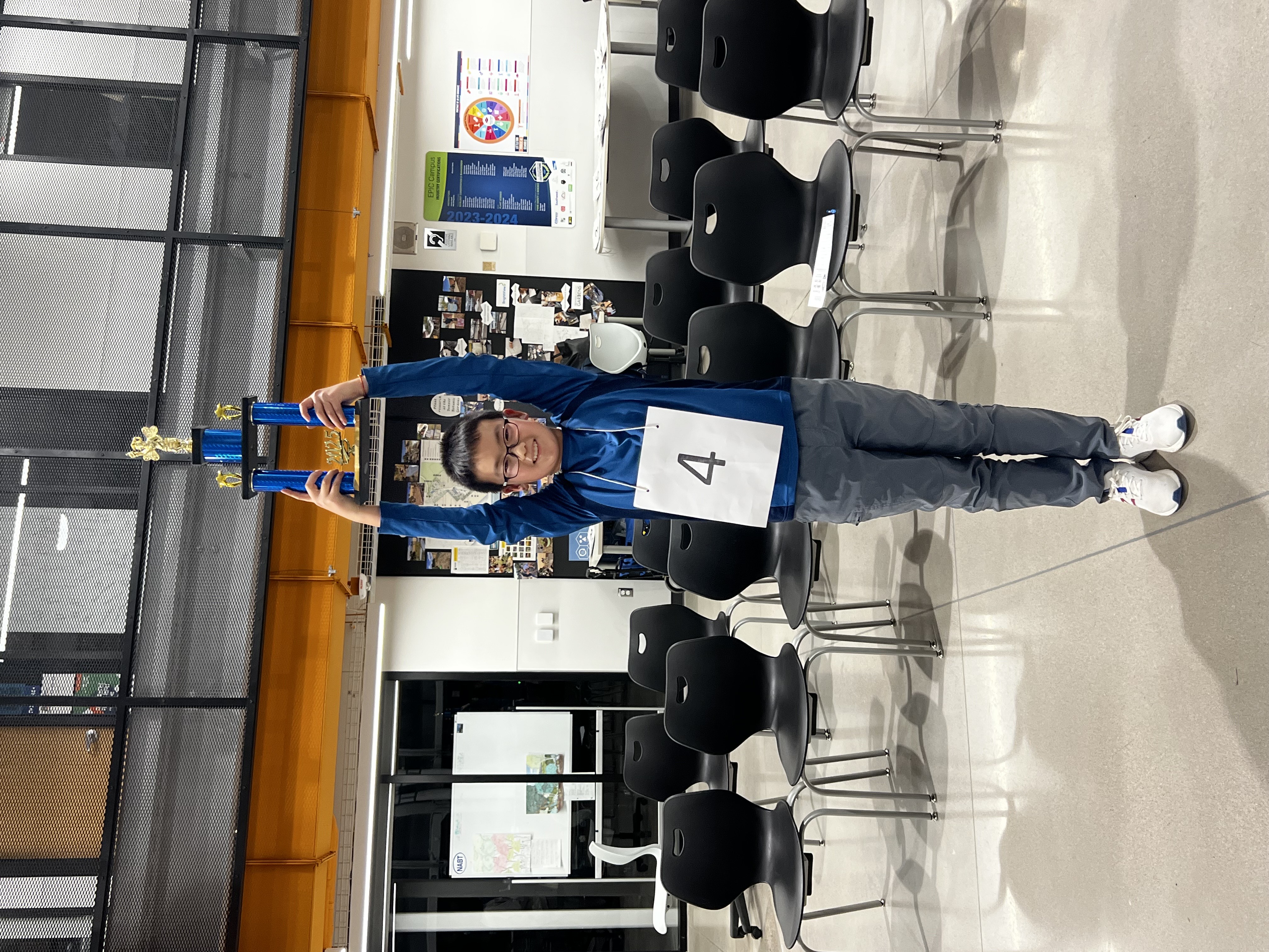 The image shows a young boy wearing glasses and a blue long-sleeved athletic jacket, standing in a modern classroom or event space. He is holding up a tall blue and gold trophy with a golden figure on top, smiling proudly. He has a white paper with the number "4" pinned to his chest, indicating participation in a competition, likely a spelling bee. The background includes black chairs arranged in rows, a whiteboard, posters, and various decorations on the walls. The setting has an industrial design with exposed metal railings and orange beams.