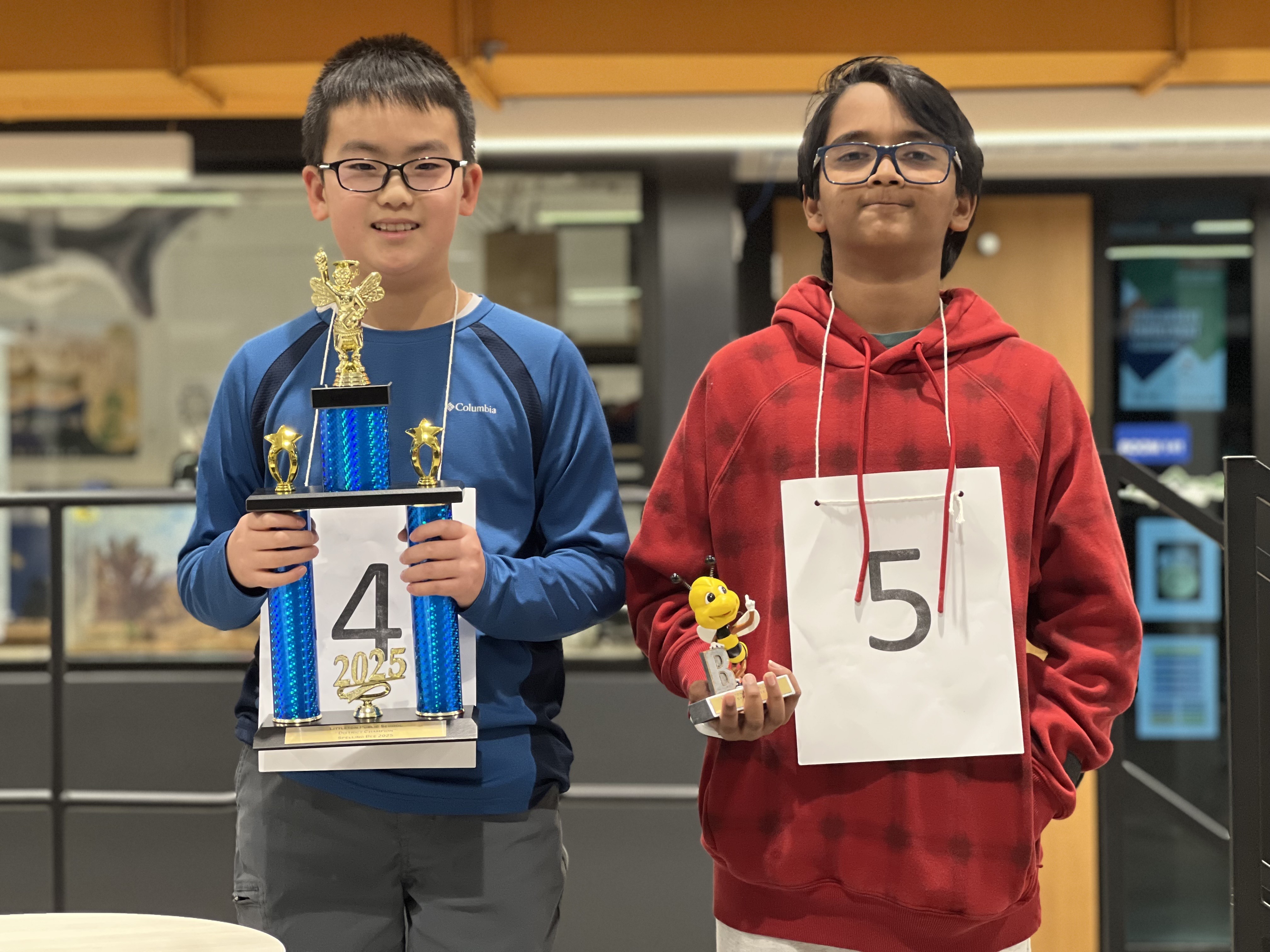 The image shows two young boys standing side by side, both wearing large white papers with numbers pinned to their chests. The boy on the left, wearing glasses and a blue long-sleeved athletic jacket, holds a large blue and gold trophy with "2025" on the base, smiling proudly. The boy on the right, wearing glasses and a red hoodie, holds a small bee-themed trophy and has a subtle smile. The background features an indoor space with modern decor, including black railings, a door, and posters on the walls. The setting appears to be a school or community event, likely a spelling bee competition.