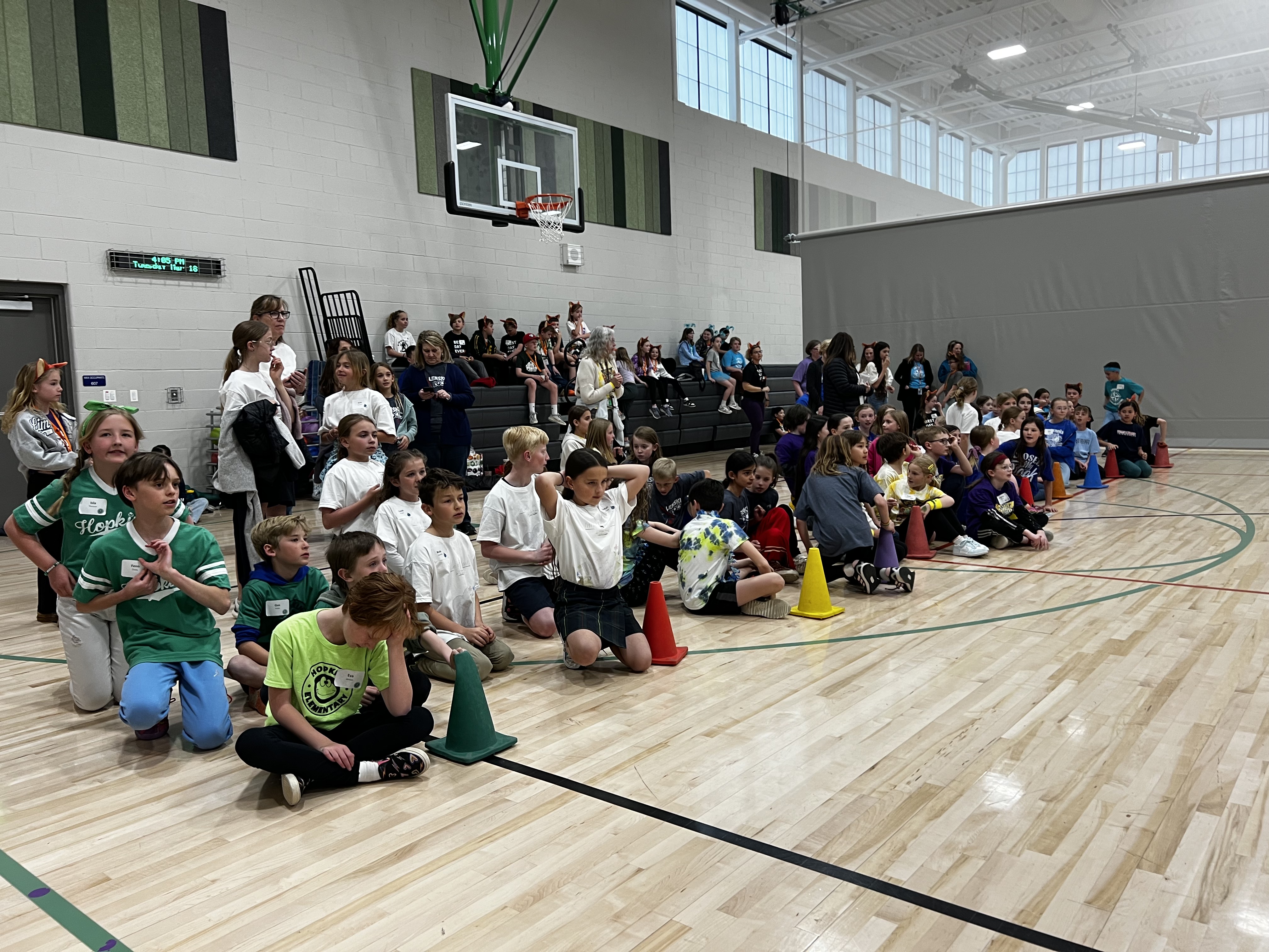 Elementary school students participate in a relay round at a Battle of the Books event in a gymnasium. Multiple teams, each wearing color-coordinated shirts, are seated or kneeling behind colored cones. Spectators, including students and adults, sit on bleachers in the background. The gym has light wood flooring, green and white wall panels, and a basketball hoop above the bleachers. The atmosphere is lively and focused.