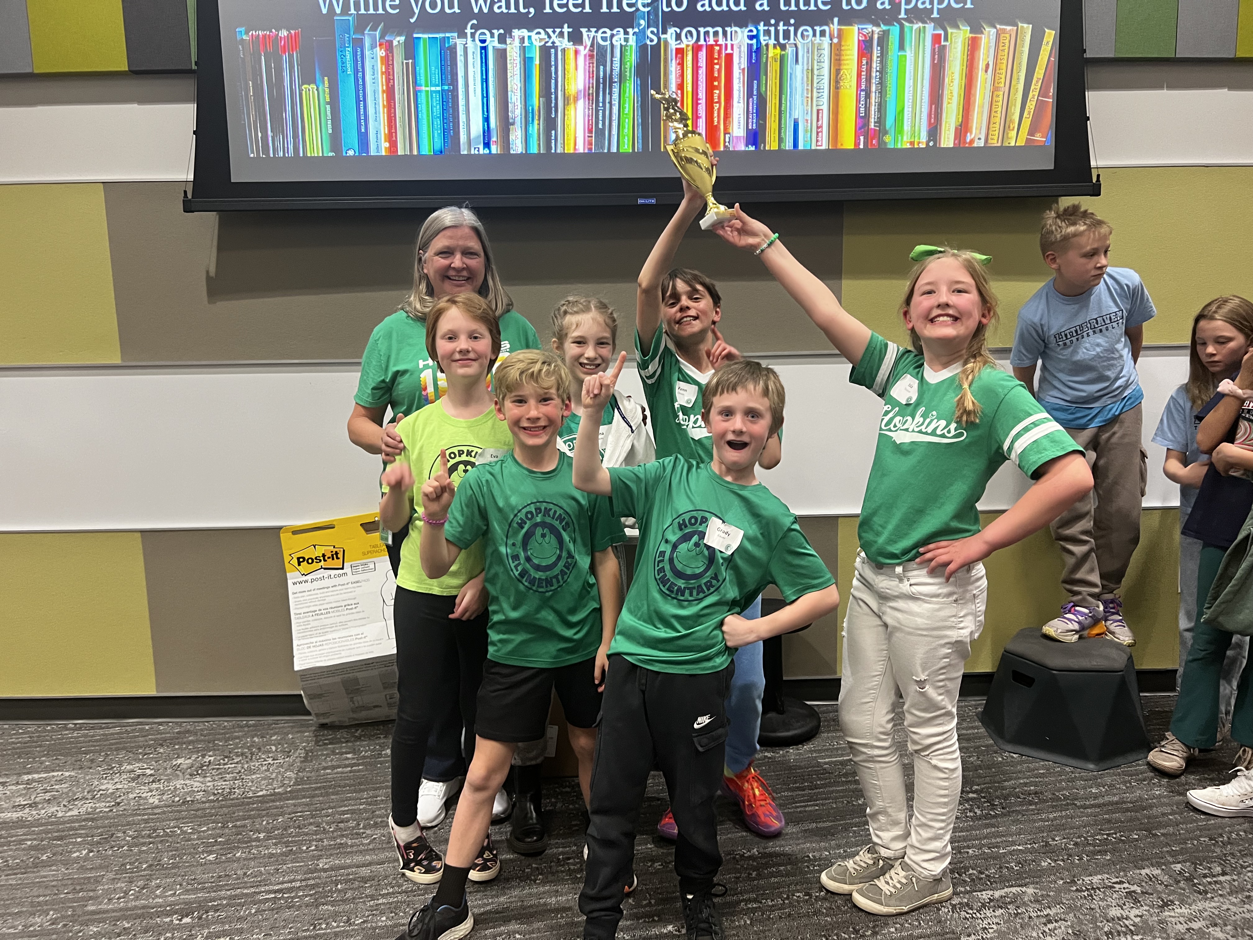 Students from Hopkins Elementary celebrate their Battle of the Books championship. Wearing green Hopkins-themed shirts, they pose proudly with a trophy held high. The group stands in front of a wall with colorful panels, smiling and making number-one gestures, while other students look on nearby.