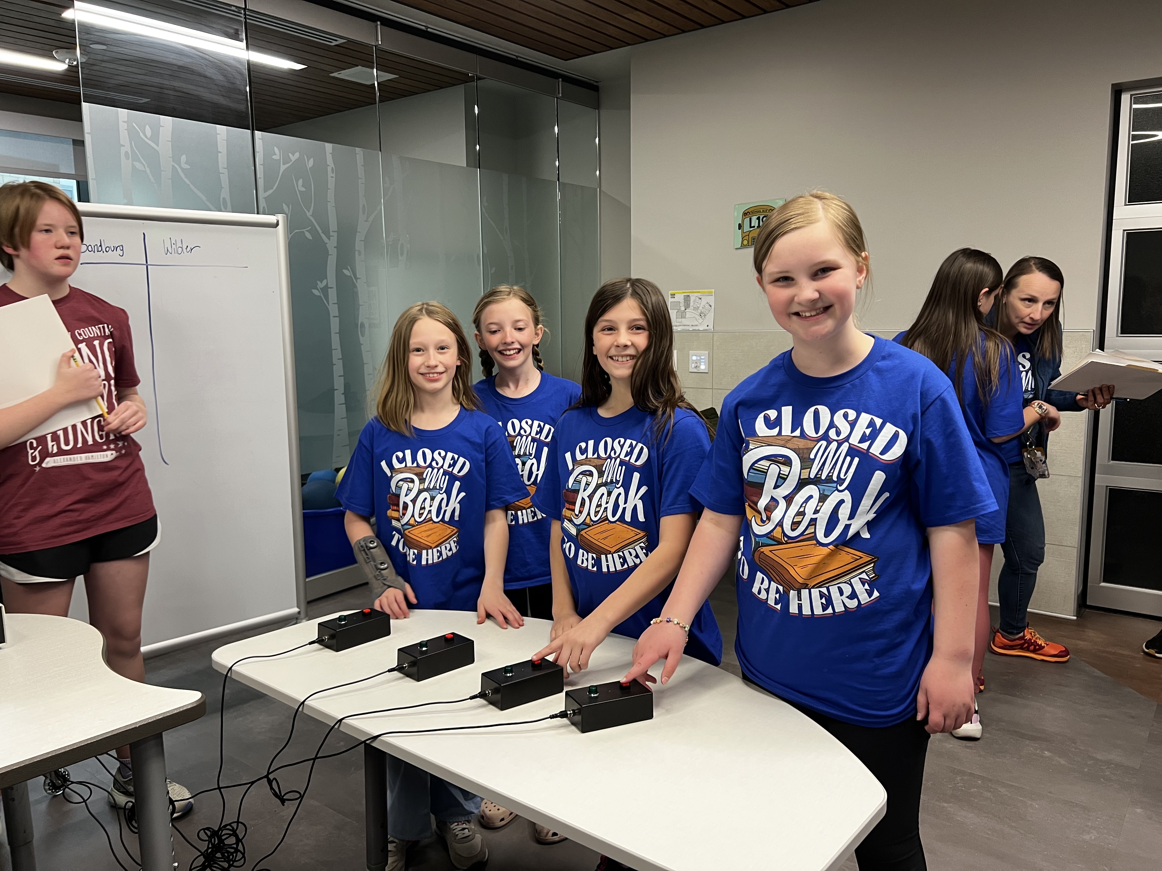Students participate in a Jeopardy-style buzzer round at the Battle of the Books event. They stand behind a table with black buzzers, smiling and ready, wearing matching blue T-shirts that read “I Closed My Book to Be Here” with an image of an open book. In the background, adults and other students are observing and taking notes, and a whiteboard displays team names.