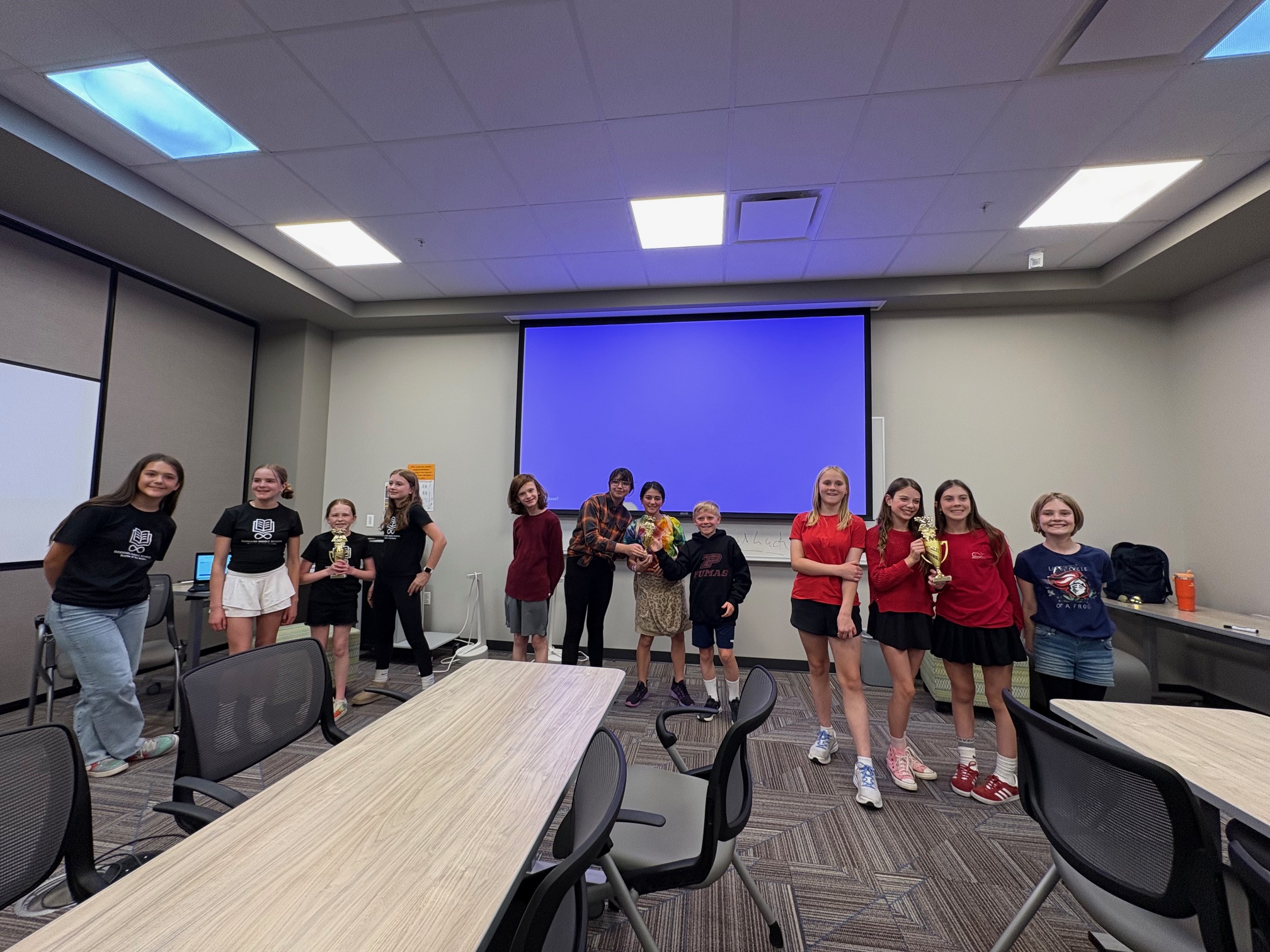 Three middle school teams pose with trophies in a classroom at a Battle of the Books event. The students are grouped in front of a large screen showing a blank blue display. Each team stands together holding a trophy, smiling, and dressed in coordinating outfits—black shirts, red tops, and casual clothing. The room has conference-style tables and chairs, and a few students and adults stand nearby.