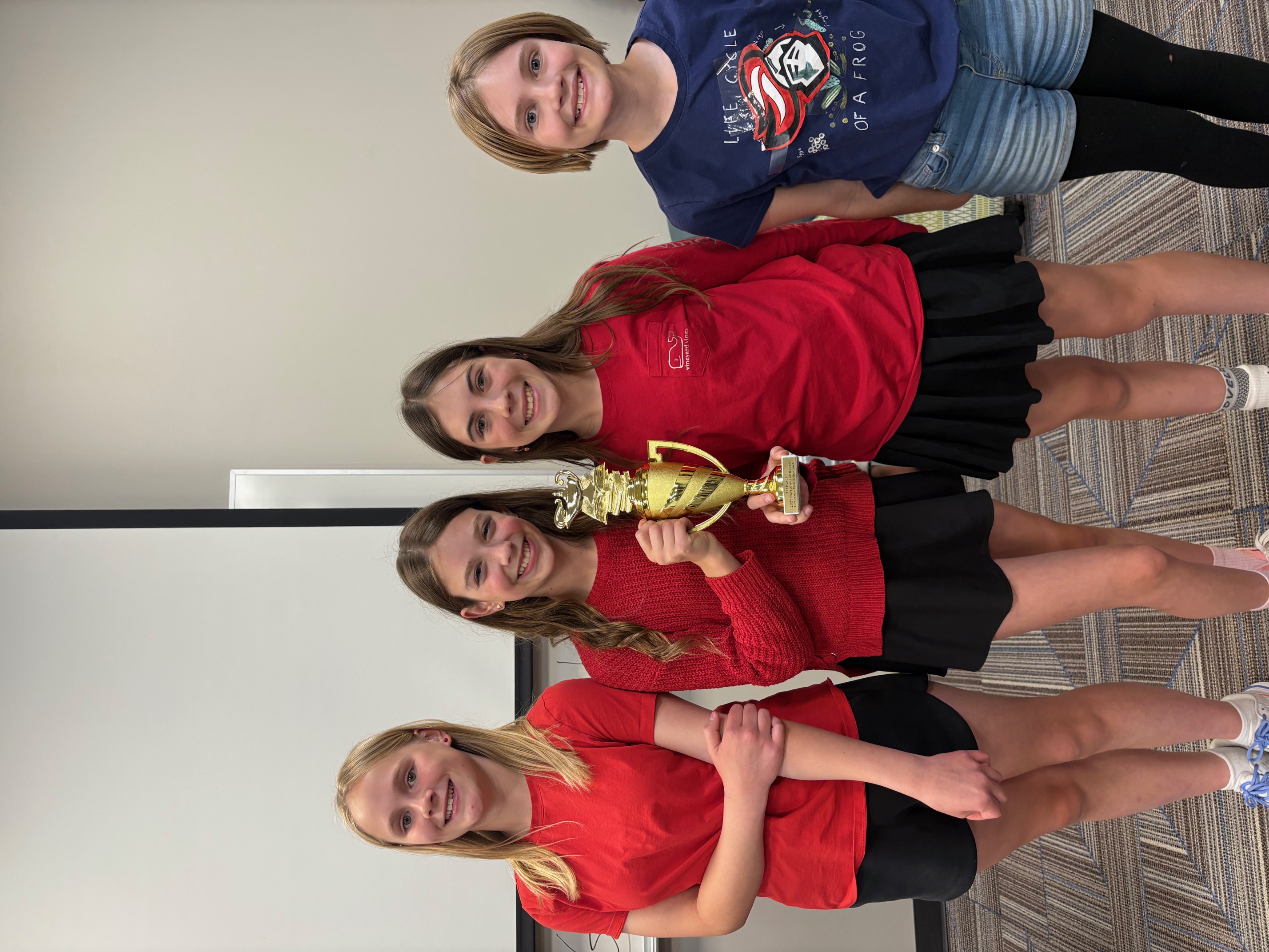 Students from Newton Middle School pose proudly with their Battle of the Books championship trophy. Wearing red and navy tops with black skirts or shorts, they smile at the camera in a classroom setting. One student holds the gold trophy at the center of the group.