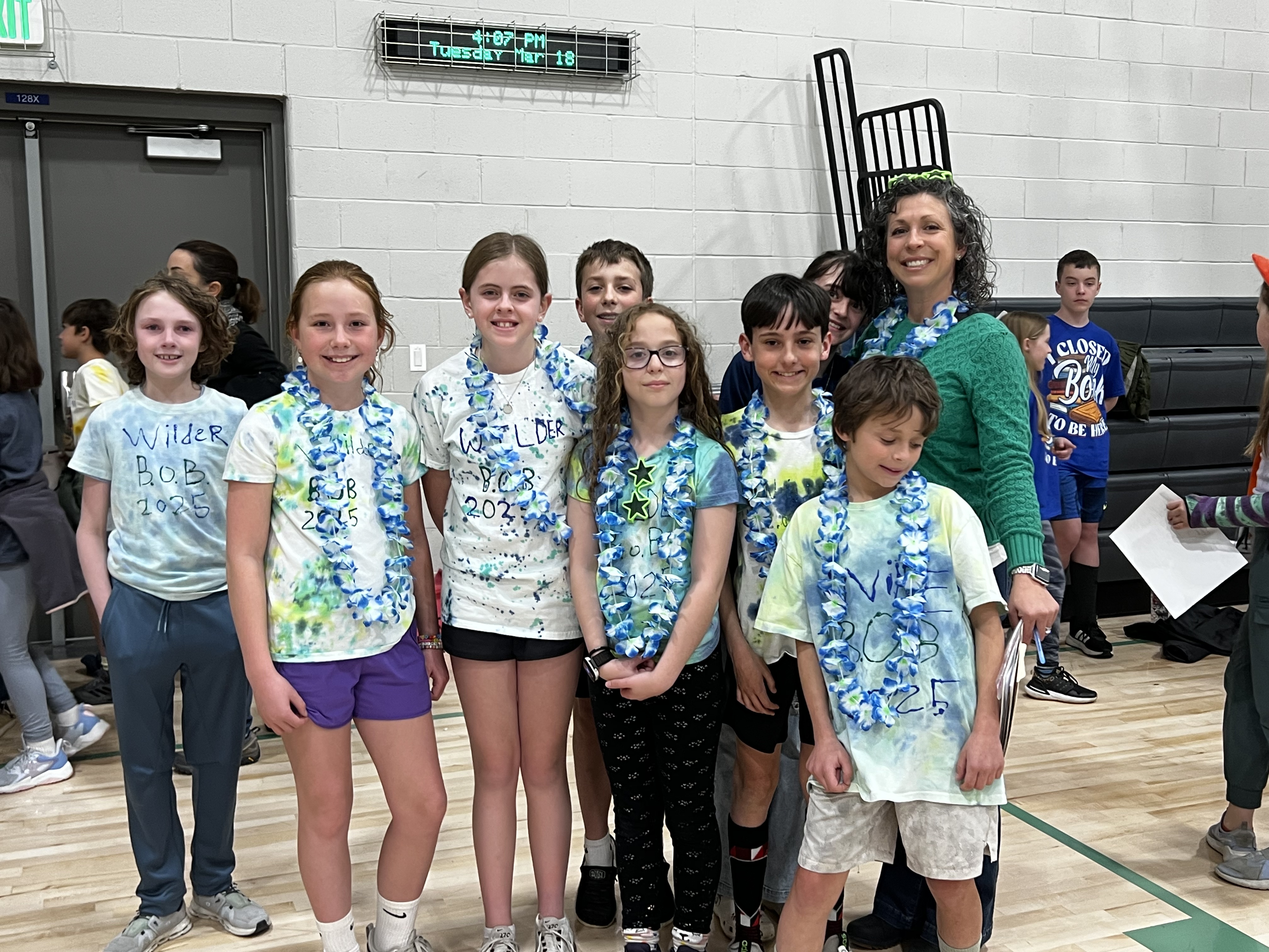 Students and an adult from Wilder Elementary pose in a gymnasium at the Battle of the Books event. The students wear matching custom tie-dye T-shirts with “Wilder B.O.B. 2025” handwritten on the front, along with blue flower leis. The adult, smiling, wears a green sweater and also has a matching lei. Other students and adults are visible in the background near the bleachers.