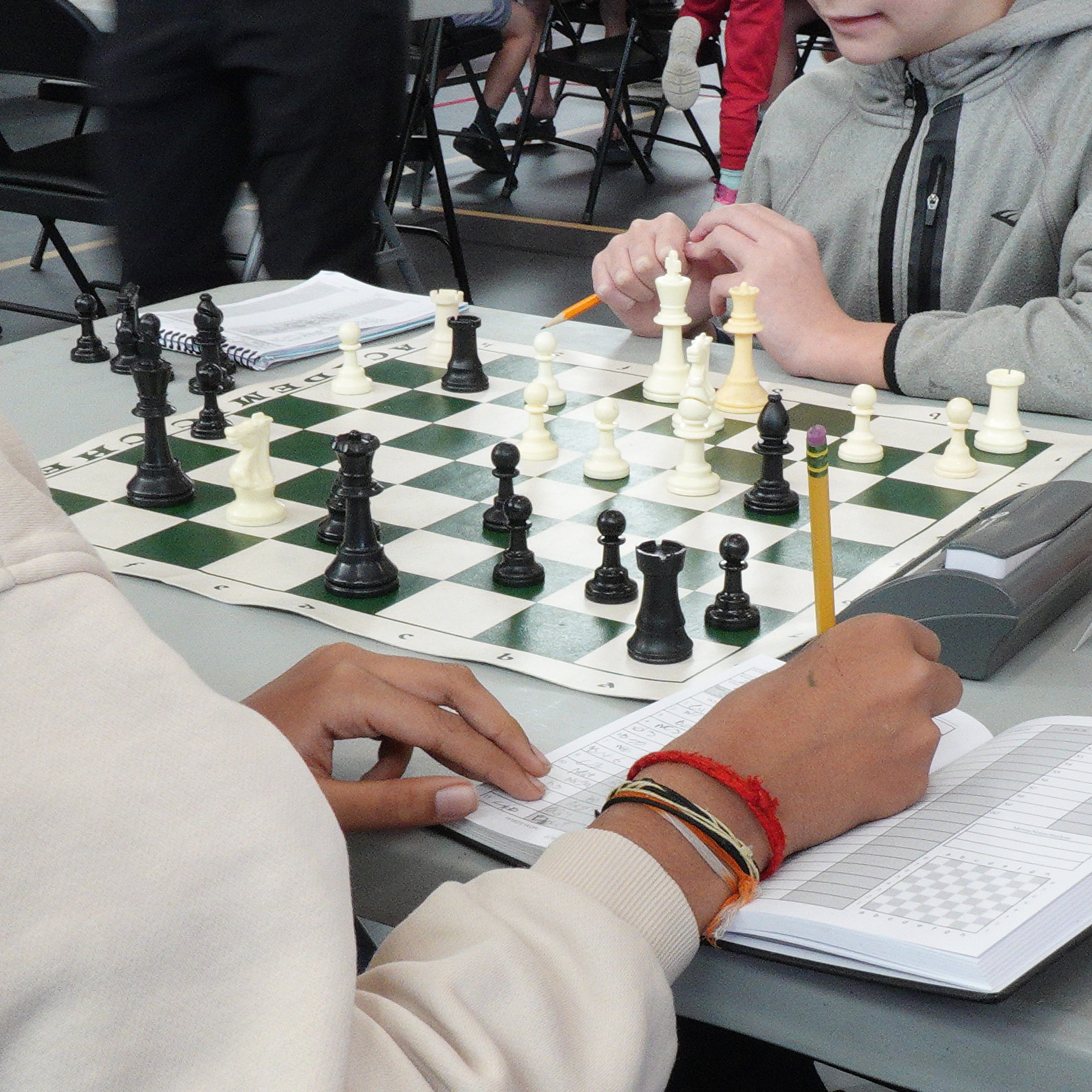 Close-up of two chess players engaged in a game, with one player taking notes on a score sheet while carefully considering their next move on a green and white chessboard.