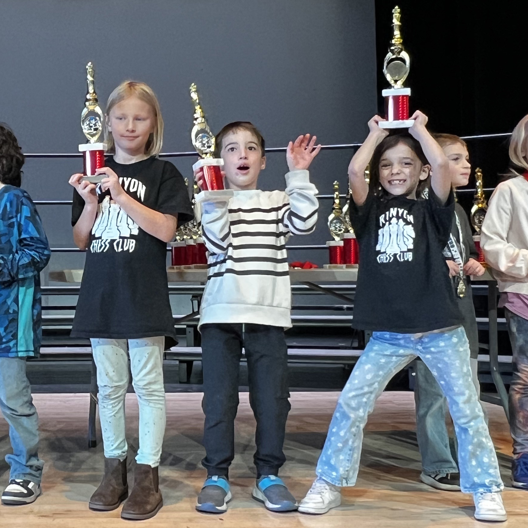 Three young chess players stand on a stage holding their red and gold chess trophies proudly, with joyful expressions and a backdrop of additional trophies on display behind them.