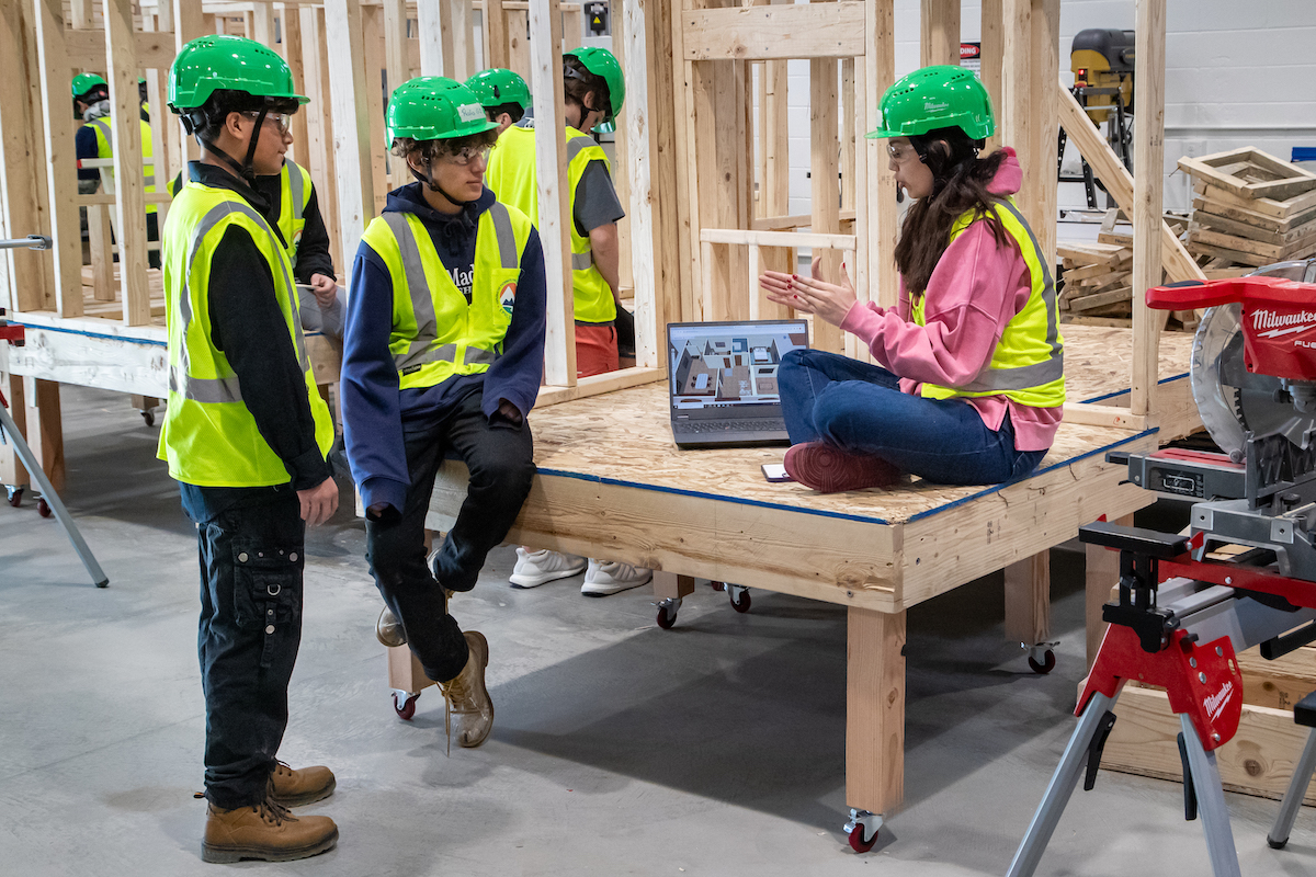 Two male students in construction PPE talk with a female student wearing PPE, who is showing them a design on a laptop. The male students are standing and the female student is sitting in a small, framed structure.