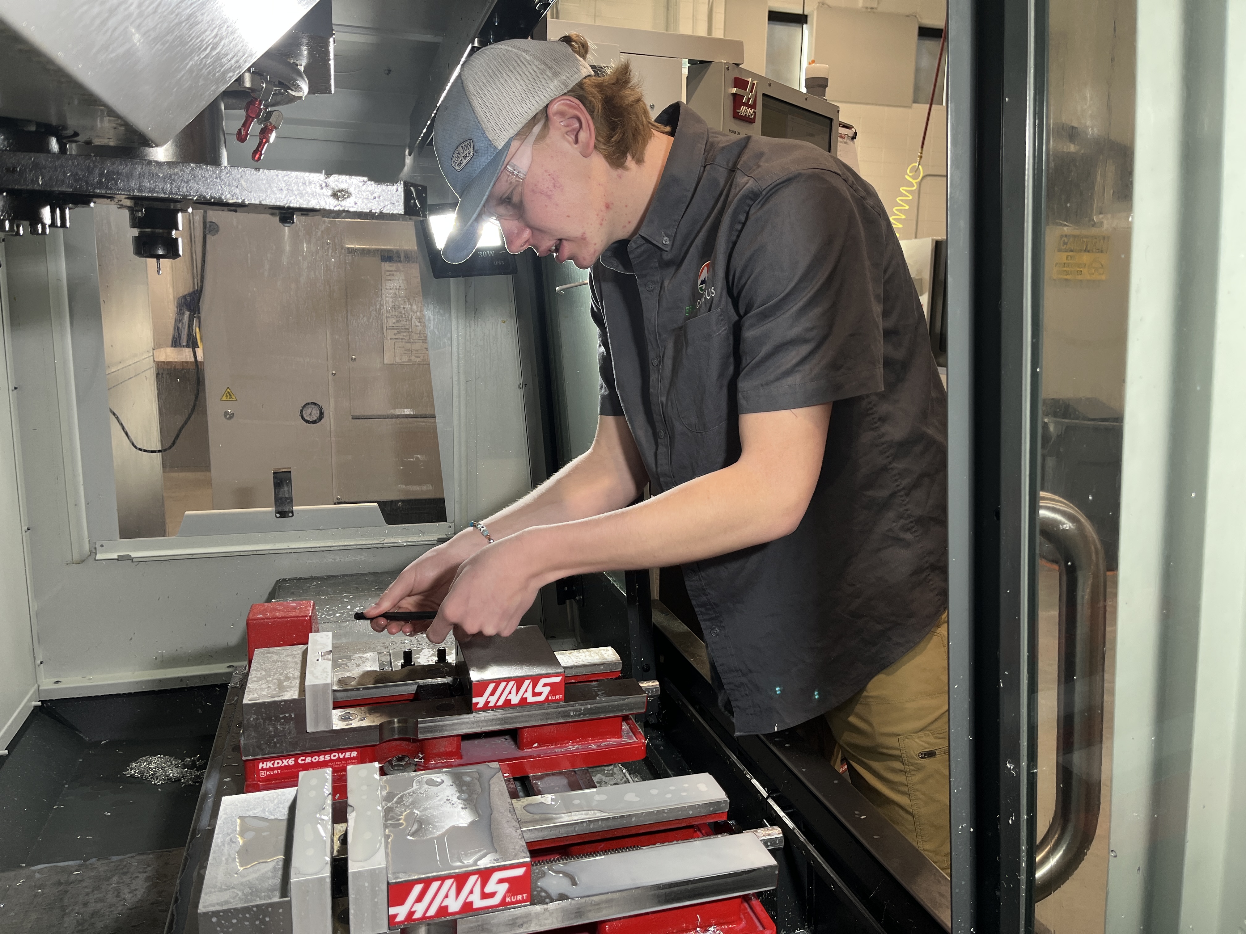 A young man wearing a gray cap, safety glasses, a dark gray work shirt with an EPIC Campus logo on the chest, and khaki pants, is working with a metal vise inside a machining area. He is adjusting a part secured in a red "HAAS" branded fixture with metal shavings visible on the surface. The background shows industrial machinery and a workspace with various equipment.