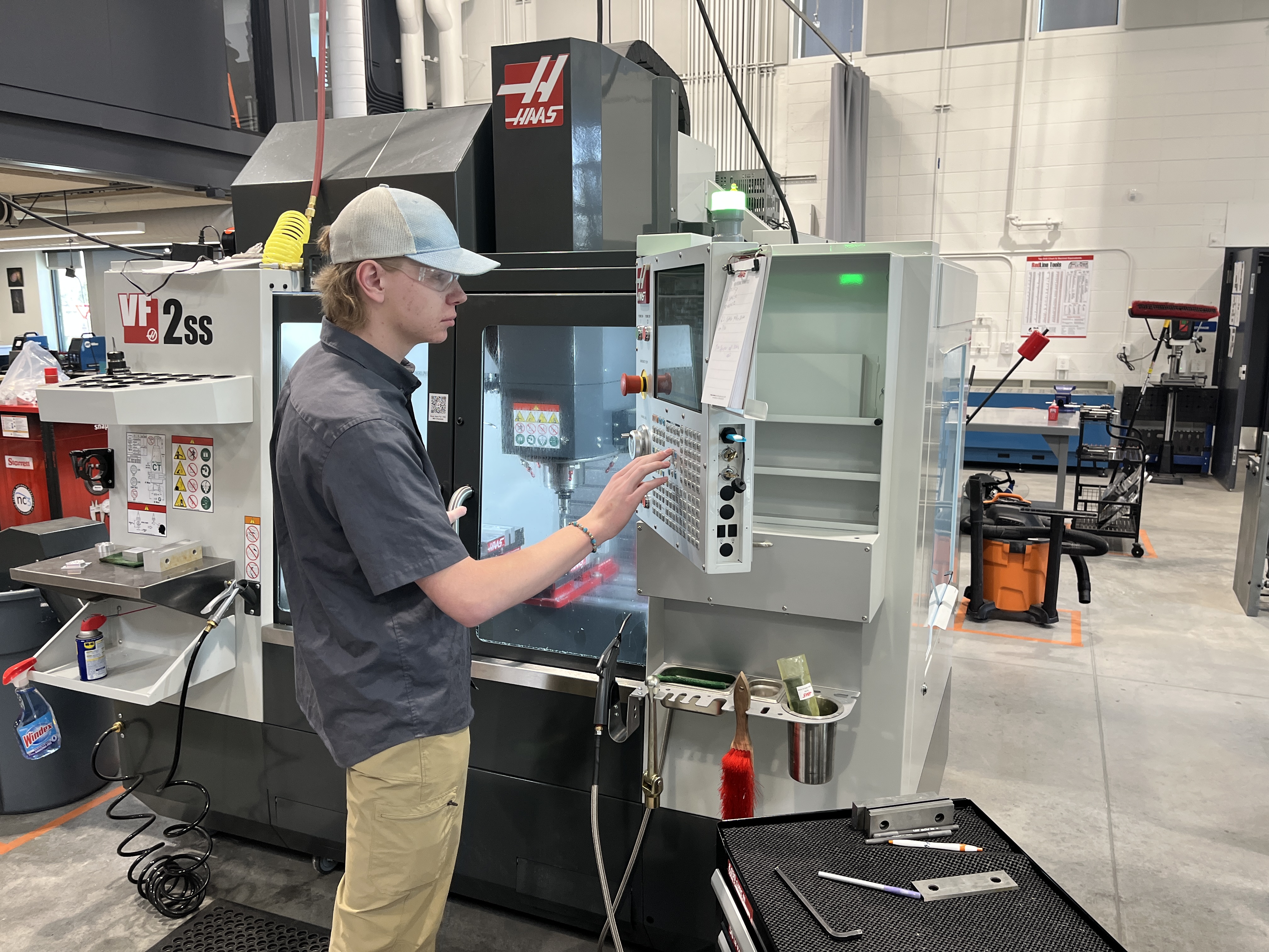 A young man wearing a gray cap, safety glasses, a dark gray work shirt, and khaki pants, is operating a HAAS VF-2SS CNC machine. He is interacting with the machine's control panel, which has a clipboard with notes attached. The CNC machine has a glass window revealing the machining area inside. The workspace is equipped with tools, cleaning supplies, and equipment such as a vacuum cleaner and tool carts in the background. The environment is clean and organized.