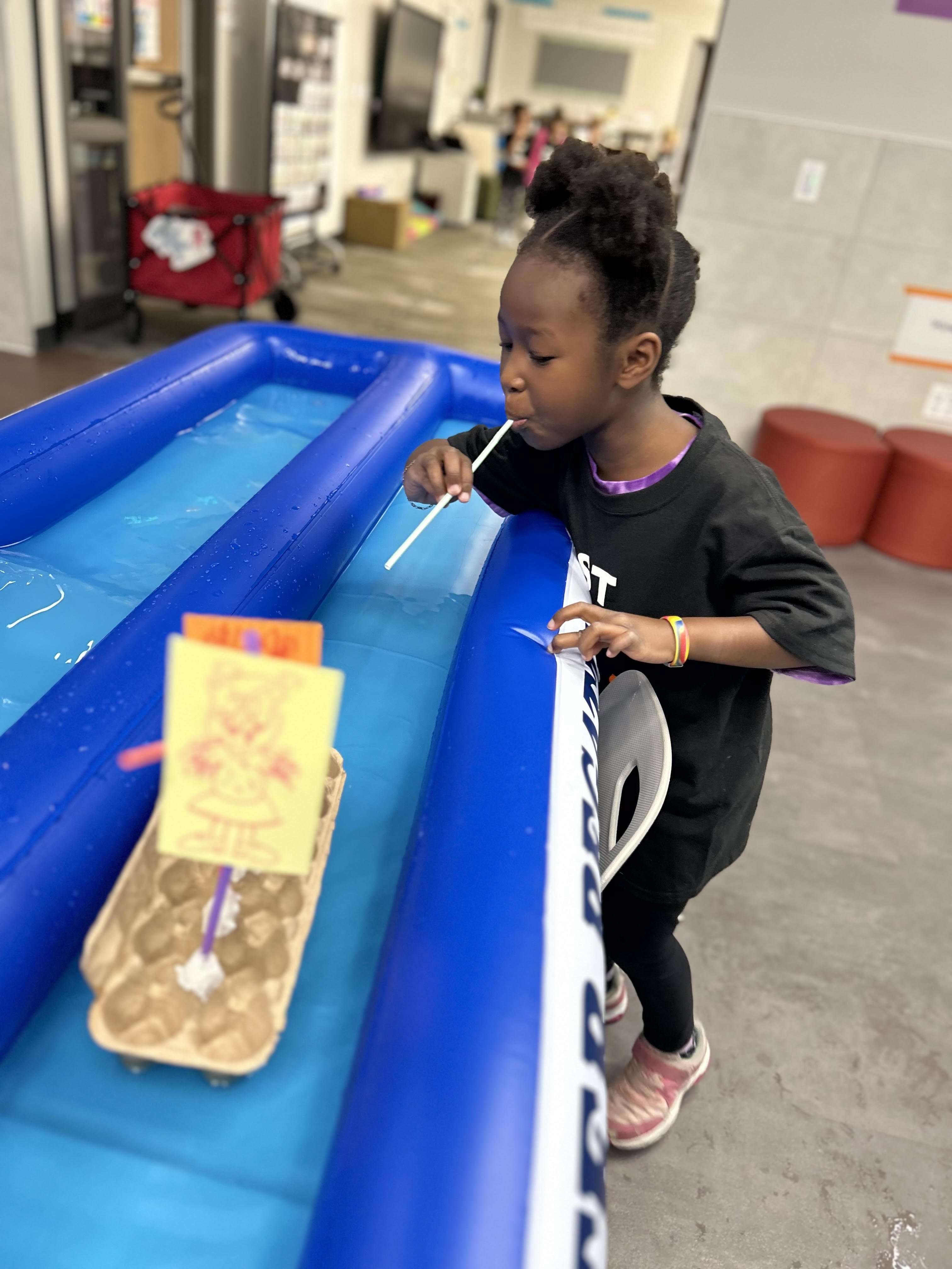 A young girl wearing a black T-shirt that says "BEST DAY EVER" leans over a blue inflatable water trough, blowing through a straw to propel a small handmade boat. The boat is crafted from an egg carton and features a yellow sail with a drawing on it. The setting is indoors, with classroom and activity supplies visible in the background. The girl is focused on the activity, participating in what appears to be a fun and creative challenge.