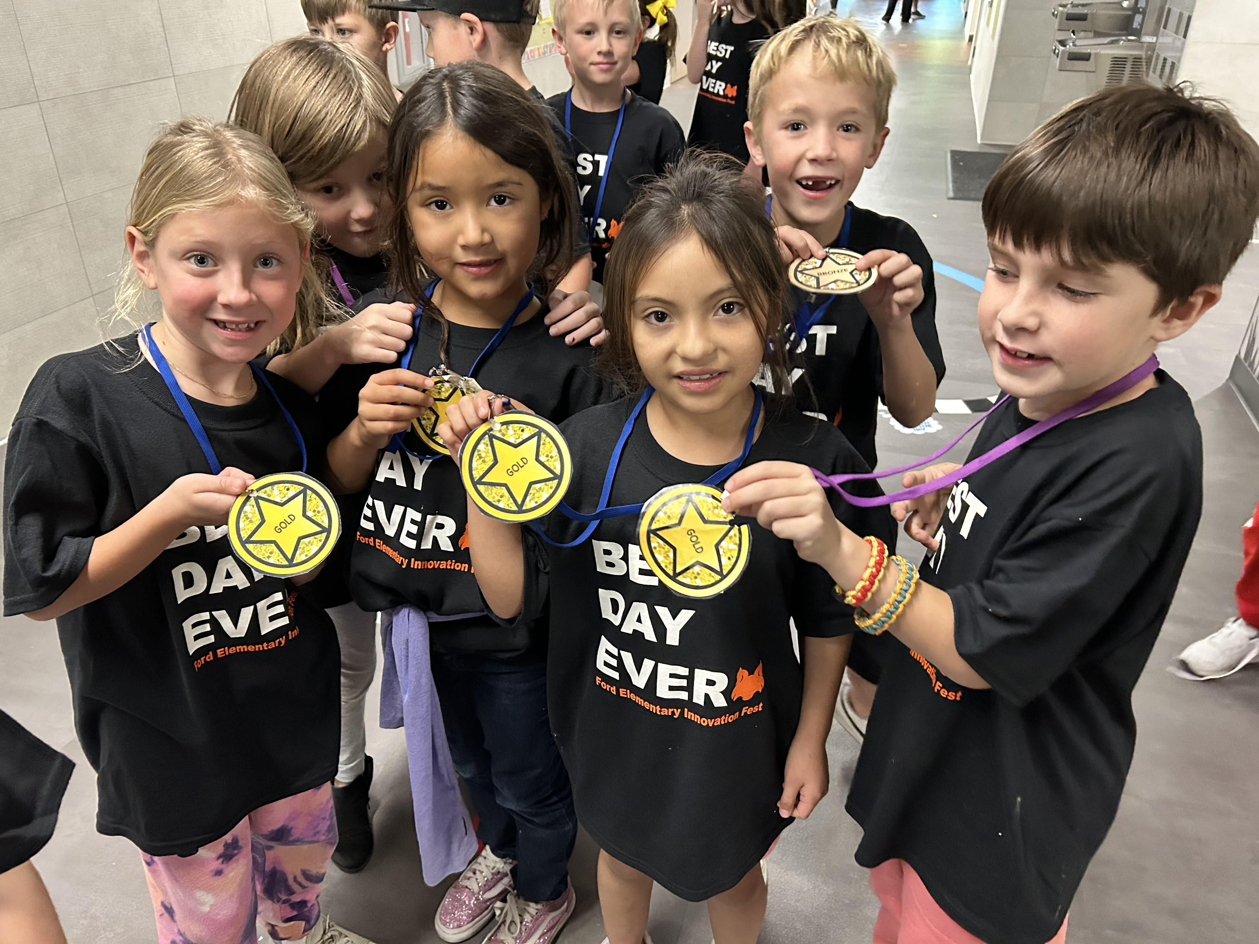 A group of smiling children wearing black T-shirts that say "BEST DAY EVER" on the front are gathered together in a brightly lit hallway. They are holding up gold star-shaped medals with "GOLD" written in the center, attached to blue or purple lanyards. The children appear excited and proud, showcasing their medals.