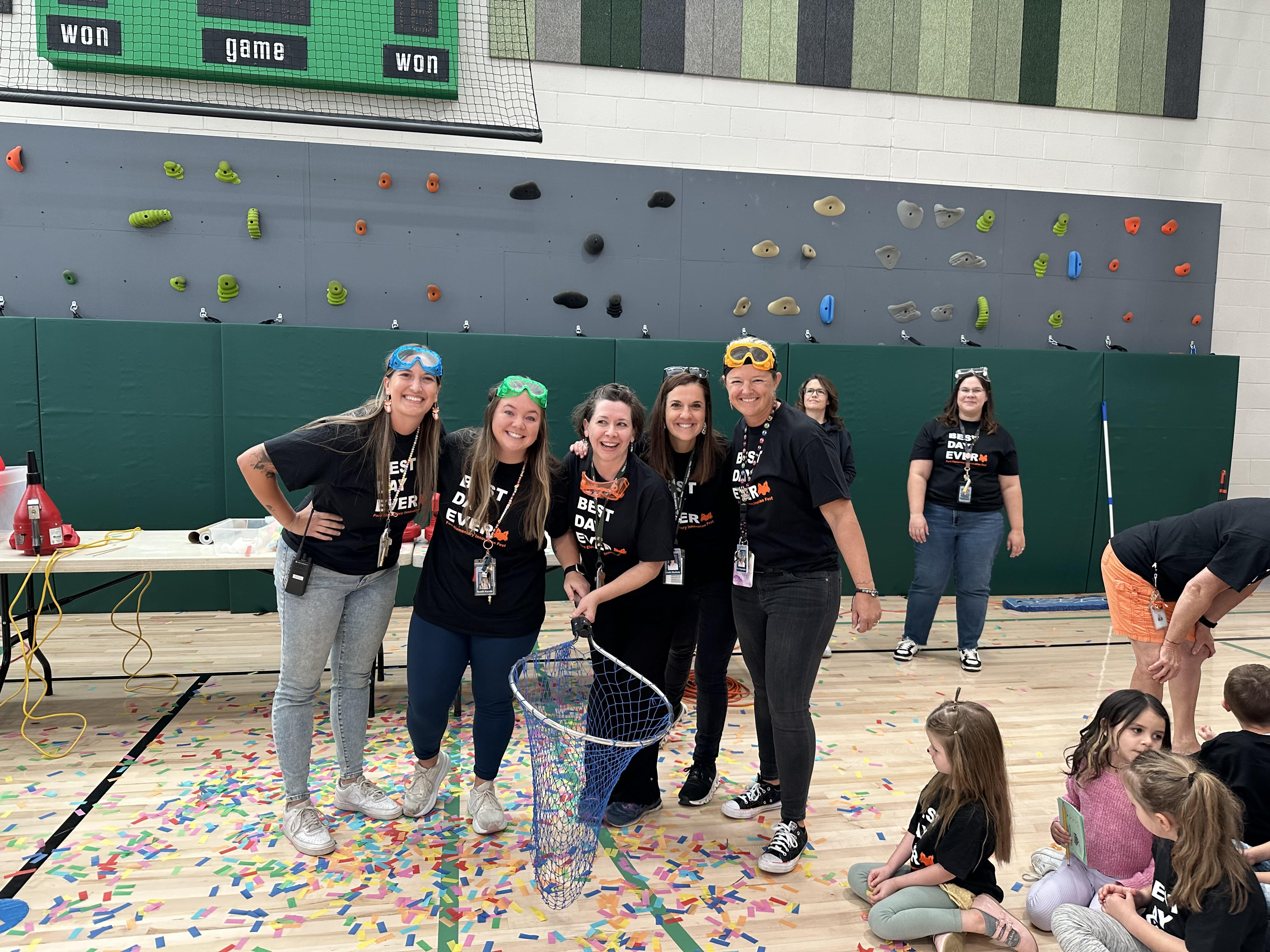 A group of five smiling adults wearing black T-shirts that say "BEST DAY EVER" pose together in a gymnasium, holding a large net. They are wearing colorful protective goggles and standing on a floor covered with multicolored confetti. In the background, there is a climbing wall with brightly colored holds and a scoreboard. Additional adults and children are visible, some sitting on the floor and participating in the event. The atmosphere is lively and festive, suggesting a school or community celebration.