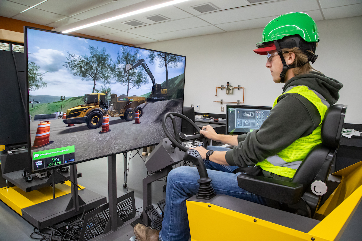 A male student wearing construction PPE is learning on a heavy equipment simulator.