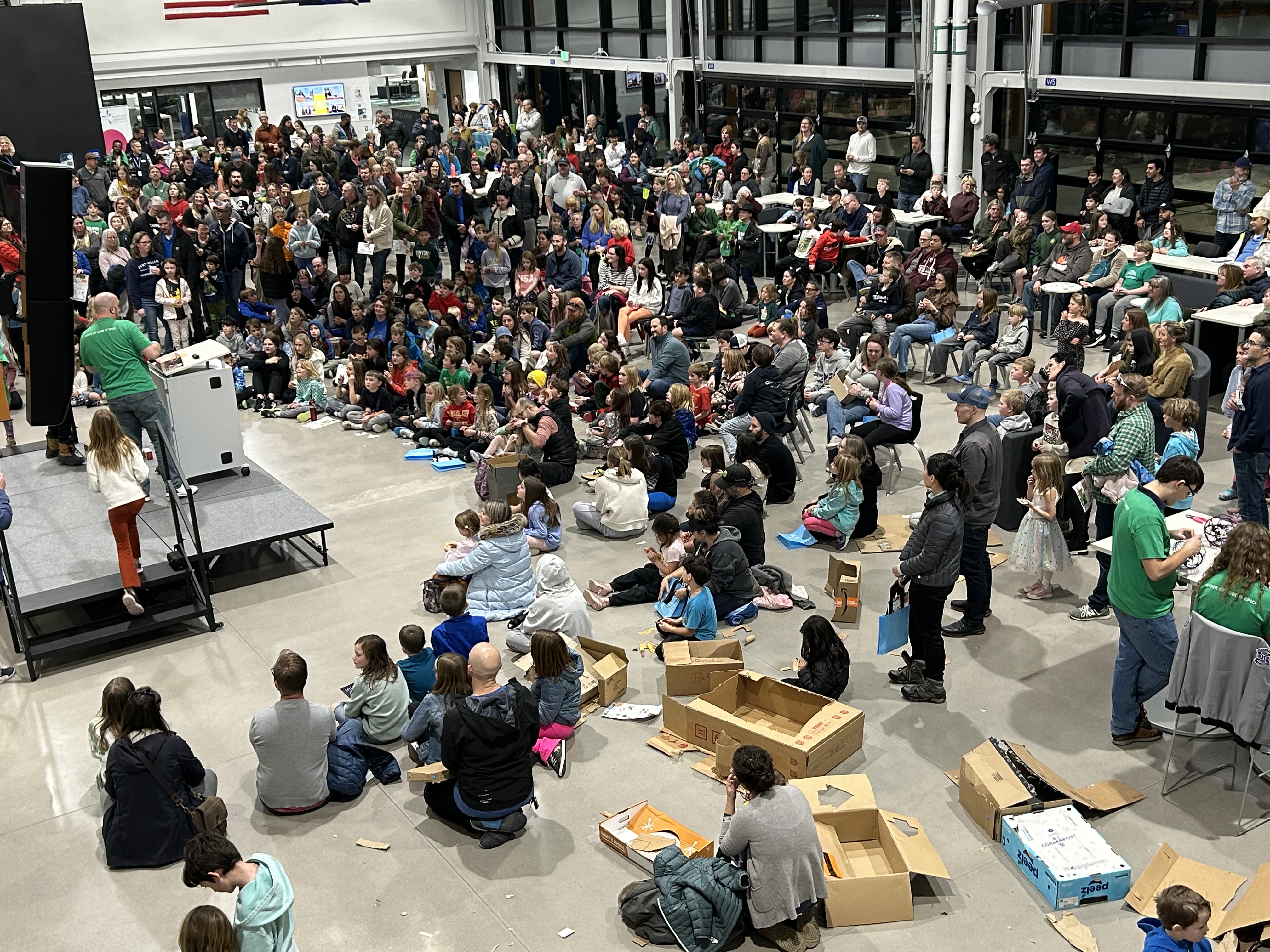 A photo taken in the EPIC Campus Great Hall looks down on hundreds of attendees facing a stage on the left side of the photos. The event coordinator, wearing a green tshirt, is speaking to the crowd from behind a podium.