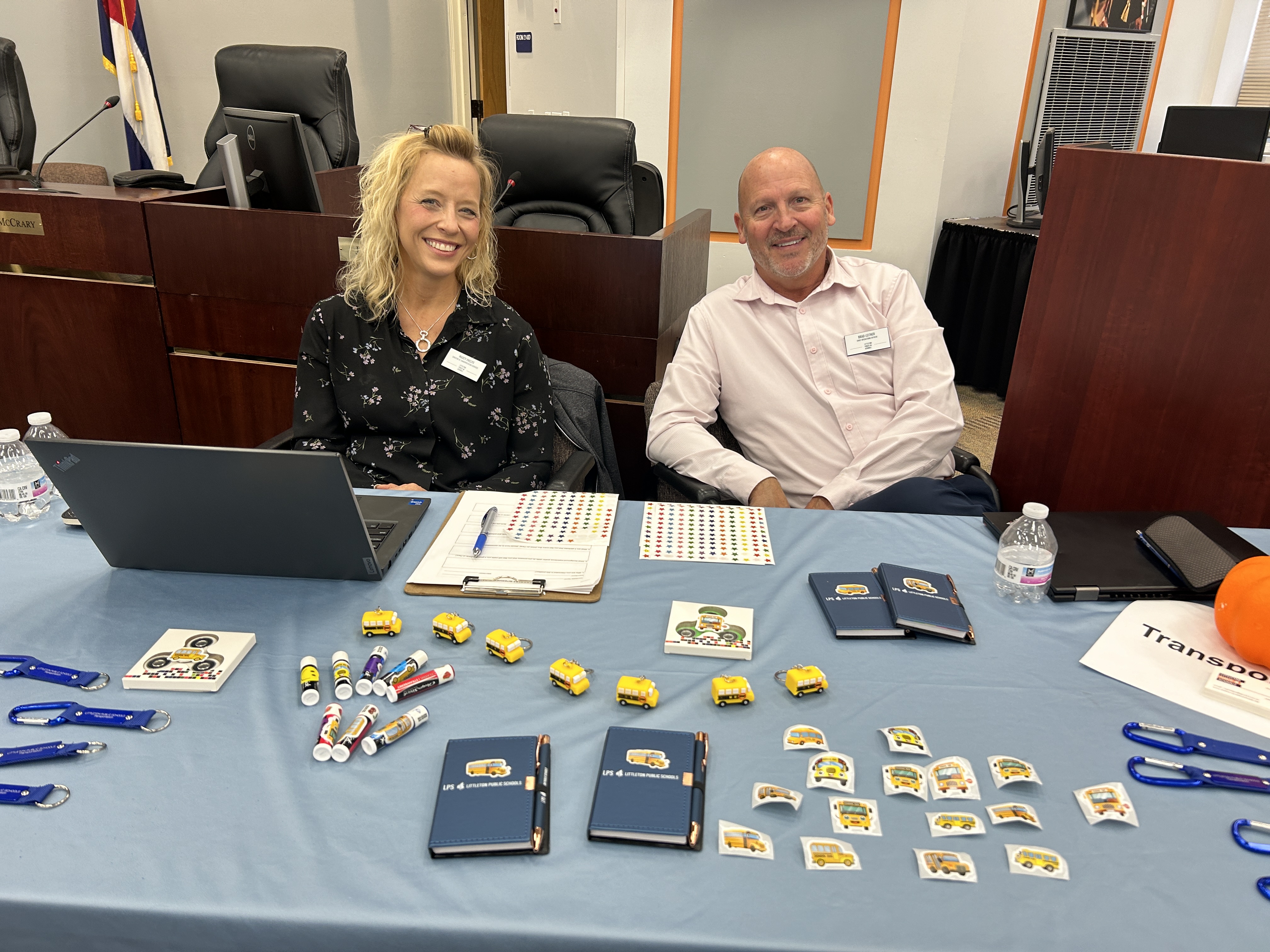 Two smiling individuals seated behind a table at a job fair event. The table is covered with a blue cloth and displays various items, including toy school buses, notebooks, pens, stickers, and lanyards. Laptops, clipboards, and water bottles are also on the table. The background features a formal meeting room setting with chairs and a Colorado flag.