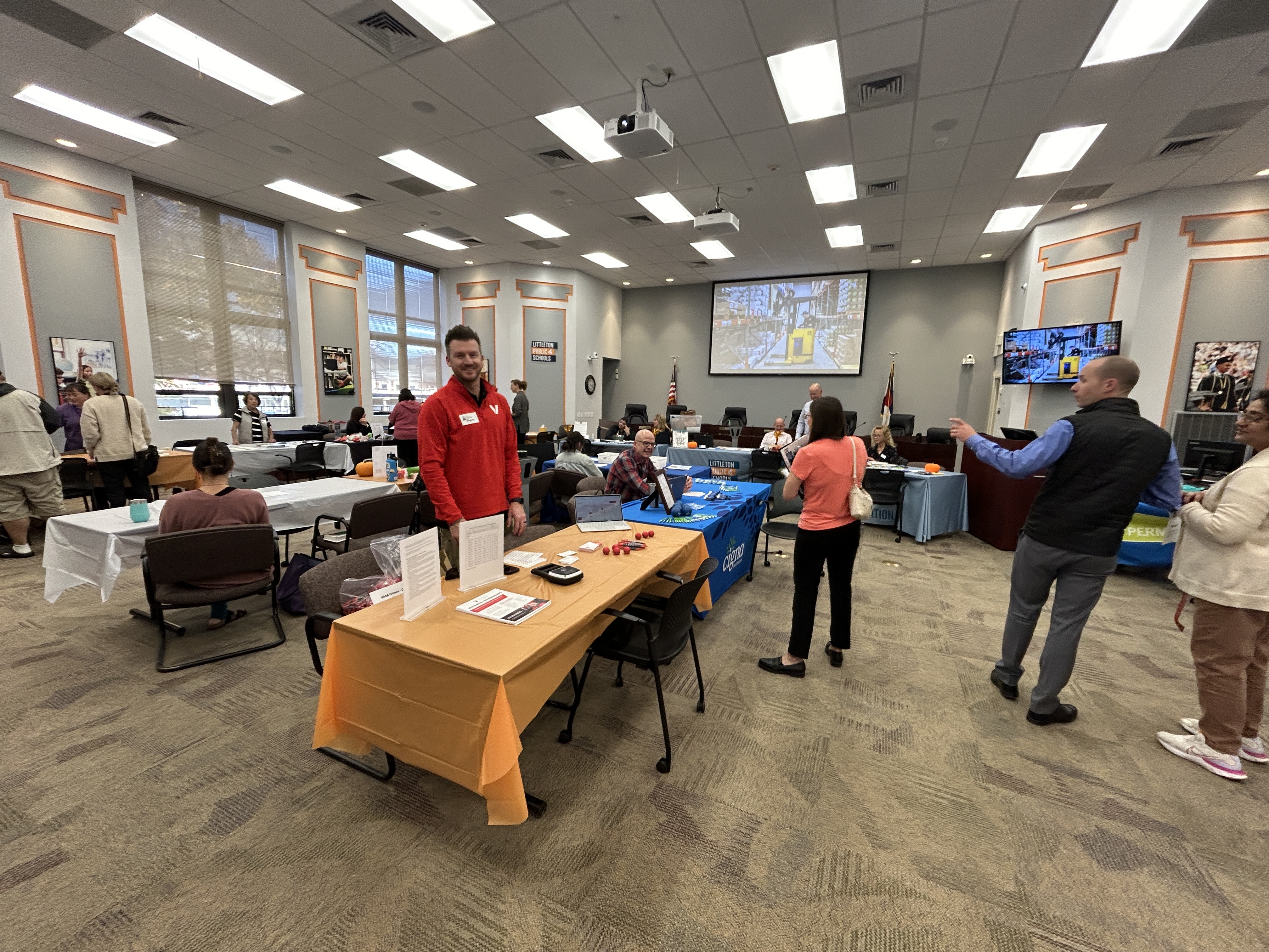 A wide view of a job fair and wellness expo held in a large, well-lit room. Several tables are set up with representatives from various organizations and LPS departments, displaying informational materials and promotional items. Attendees are engaging with exhibitors, while a person in a red shirt stands near a table with an orange tablecloth, smiling at the camera. Large screens and American flags are visible in the background, emphasizing the formal setting.