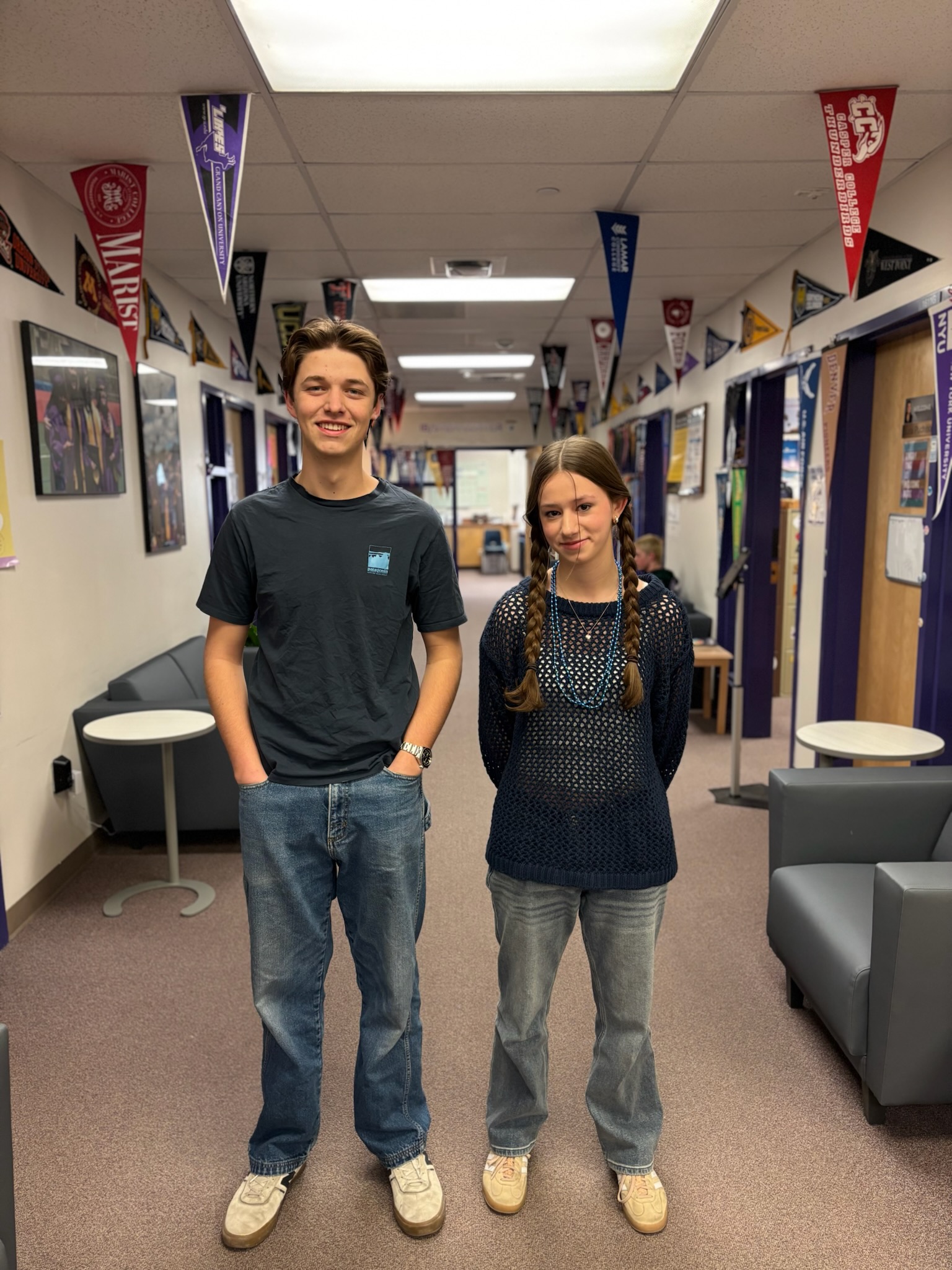 A young man and a young woman stand side by side in a school hallway, both smiling at the camera. The young man on the left wears a dark blue t-shirt with a small logo, blue jeans, and beige sneakers. He has short, wavy brown hair. The young woman on the right wears a navy blue mesh sweater over a dark top, blue jeans, and beige sneakers. She has long brown hair styled in two braids and wears a beaded necklace. The hallway behind them has college pennants hanging from the ceiling, purple doors, framed pictures on the walls, and gray seating areas with small round tables.