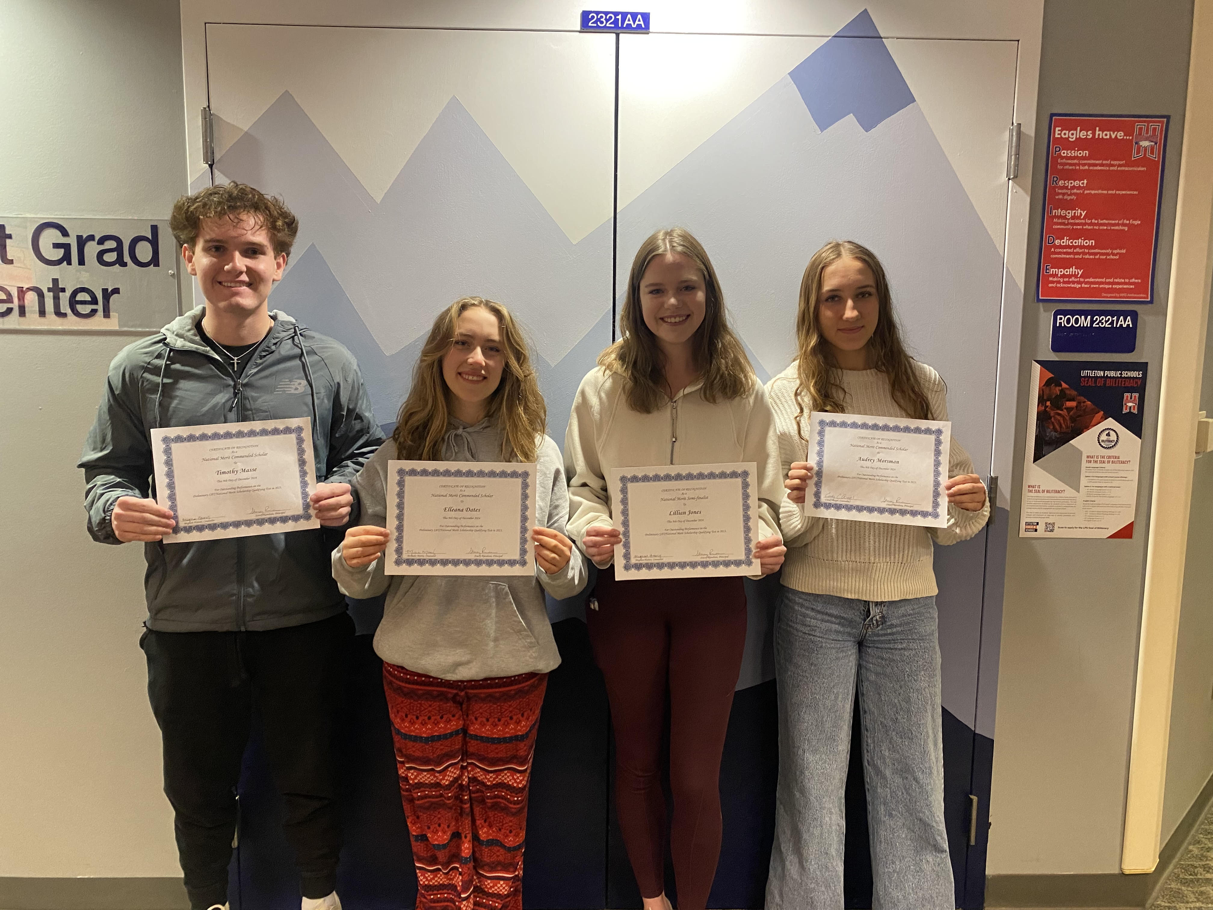 Four high school students stand side by side in front of a blue and white geometric-patterned background, holding certificates. The group includes one young man on the left and three young women, all smiling at the camera. The young man wears a dark gray athletic jacket, black pants, and a cross necklace. The first young woman wears a gray hoodie and red patterned pants. The second young woman wears a white zip-up sweater and maroon leggings. The third young woman wears a white knit sweater and light blue wide-leg jeans. To the right, a red and white poster displays values such as 'Passion,' 'Respect,' and 'Integrity,' along with a 'Littleton Public Schools' Seal of Biliteracy flyer. The setting appears to be a school hallway or office.