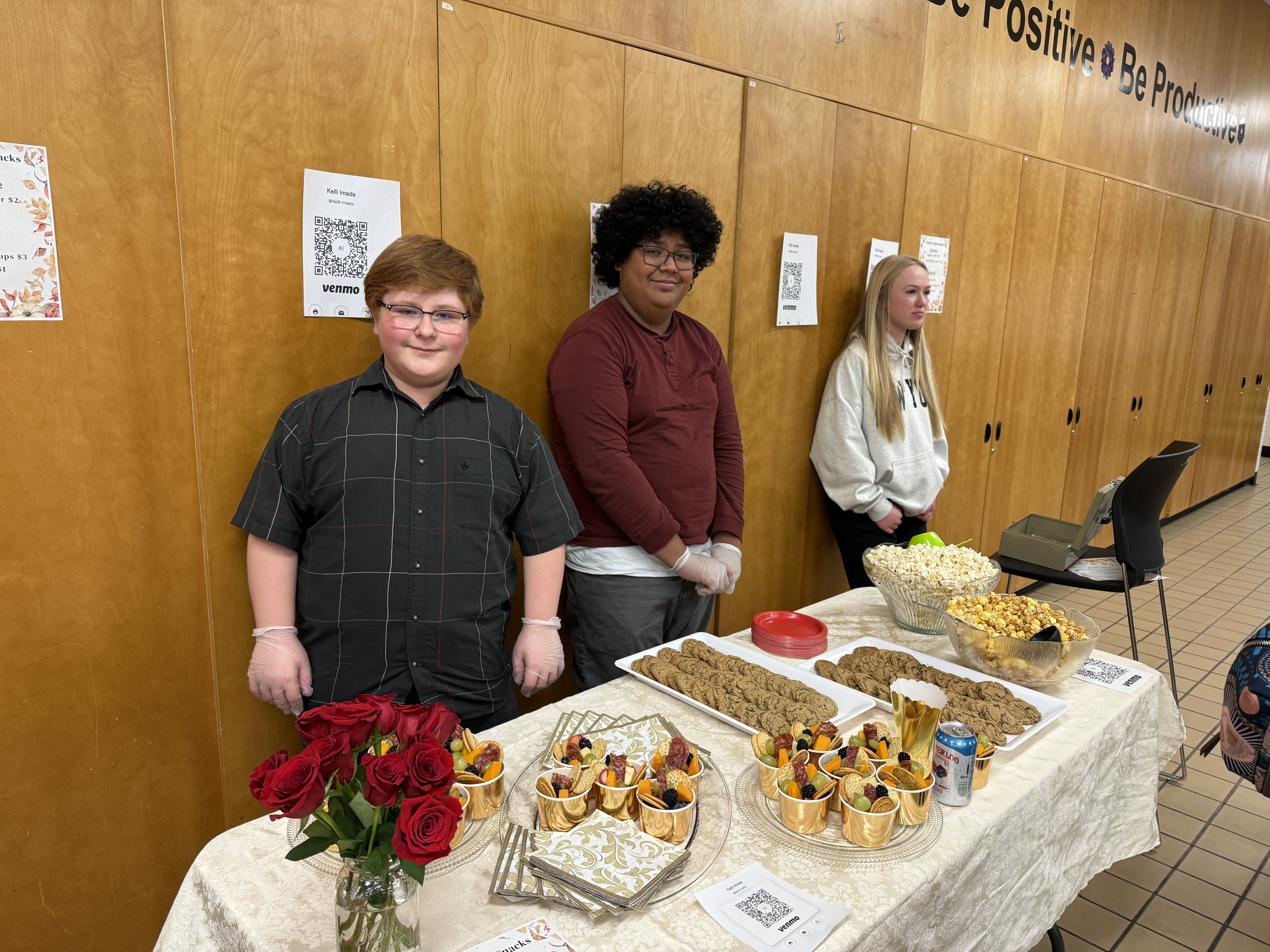 Three students stand behind a snack table at an event, with a light-colored cloth covering the table. The table displays a variety of snacks, including bowls of popcorn, plates of cookies, and gold cups filled with fruit. A vase of red roses decorates the table. The students, two wearing gloves, smile while standing in front of a wooden wall with QR codes for payment options visible. The event appears organized and inviting, suggesting a school or community gathering.