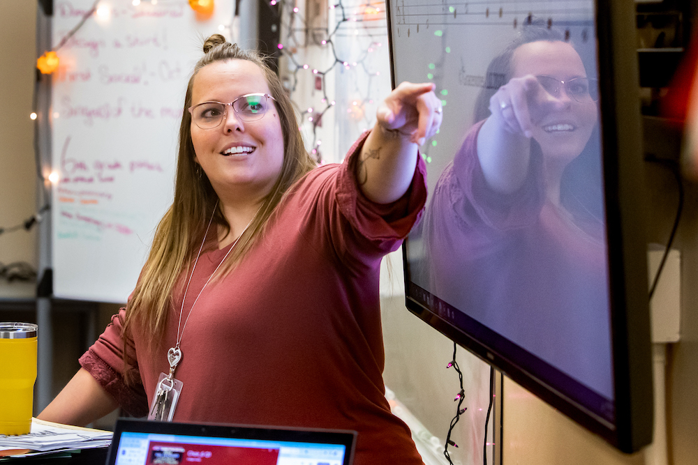Female teacher wearing glasses and a red shirt gestures to her left to a wall-mounted screen.