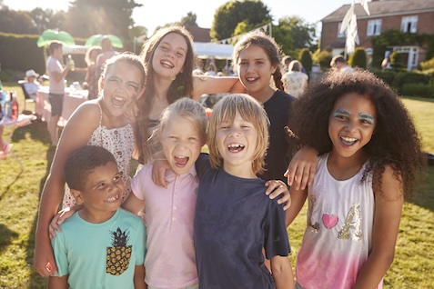 Group of seven smiling children of diverse backgrounds standing close together outdoors at a sunny event. They are posing for the camera with their arms around each other. The setting appears to be a backyard or park with other people and activities visible in the background.