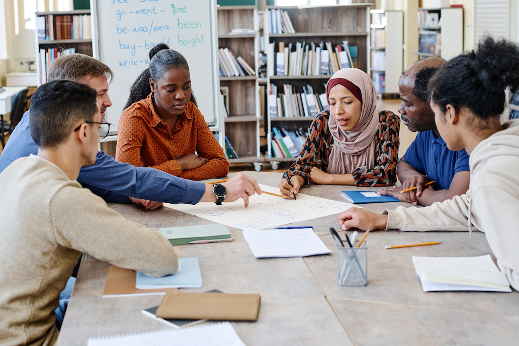 A committee of diverse adults sits around a table discussing a document in a school library.