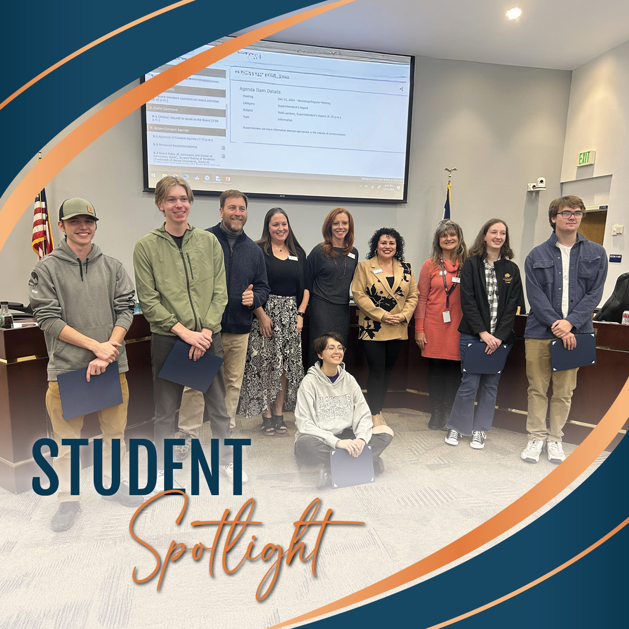 Five students holding certificates pose in front of the Board of Education members in a meeting room, with one student sitting cross-legged in the front. Everyone is smiling, and one Board member gives a thumbs-up. The image is framed with curved blue and orange graphics, with the text 'STUDENT Spotlight' in the bottom left corner.