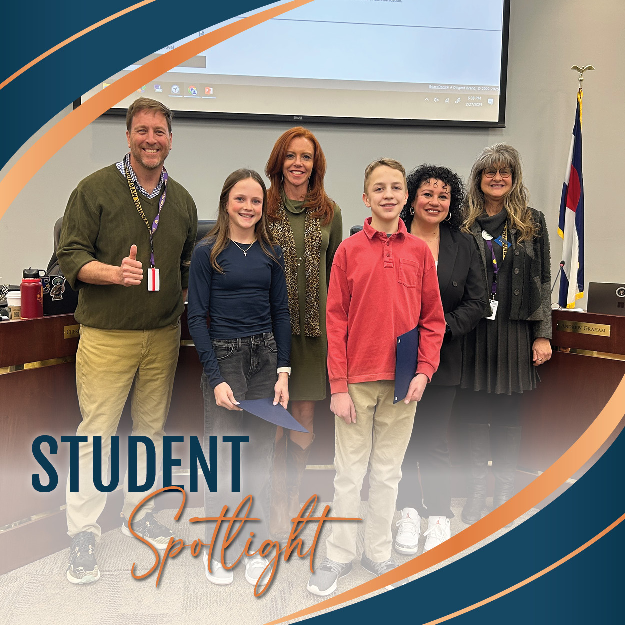 Two students stand with the Board of Education in the boardroom. The students, a girl in a navy blue top and jeans, and a boy in a red shirt and khaki pants, are holding certificates and smiling. The adults are dressed professionally, with one man giving a thumbs-up. The background shows a projector screen and a Colorado state flag. The image includes a graphic overlay with the words "STUDENT Spotlight" in bold blue and orange text, surrounded by curved design elements in blue and orange.