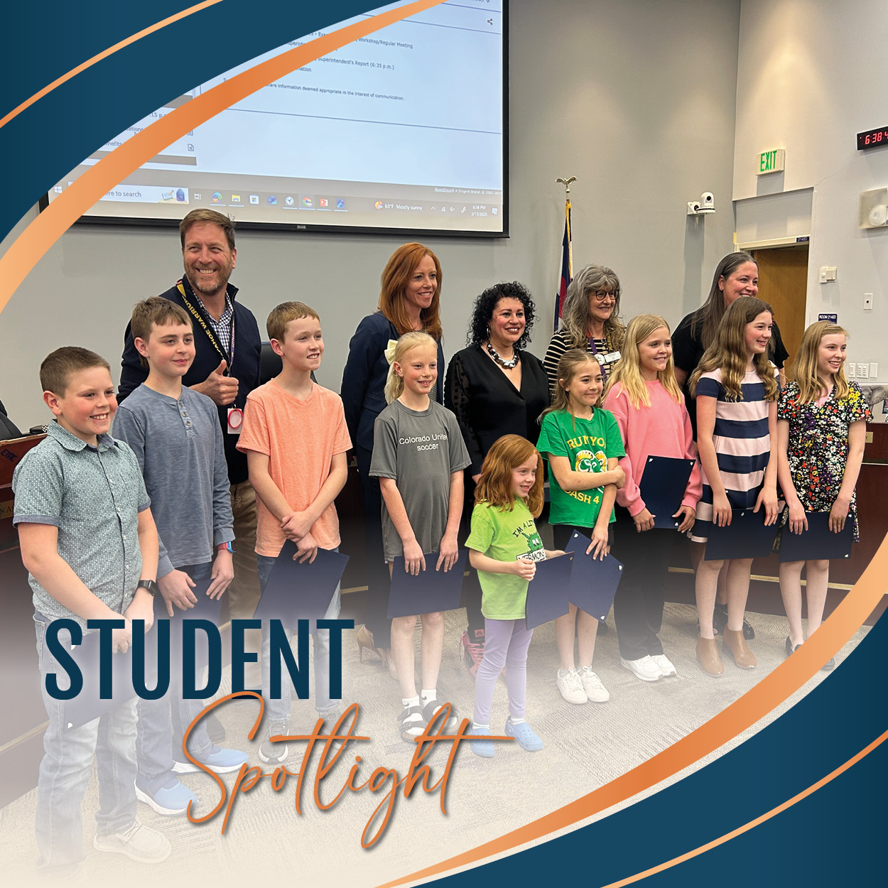 A group of 9 students stands with the Board of Education in the boardroom. The students, dressed casually in t-shirts, jeans, and casual dresses, are holding certificates and smiling. The adults, dressed professionally, are standing behind them and smiling, with one man giving a thumbs-up. The background shows a projector screen and a Colorado state flag. The image includes a graphic overlay with the words "STUDENT Spotlight" in bold blue and orange text, surrounded by curved design elements in blue and orange.