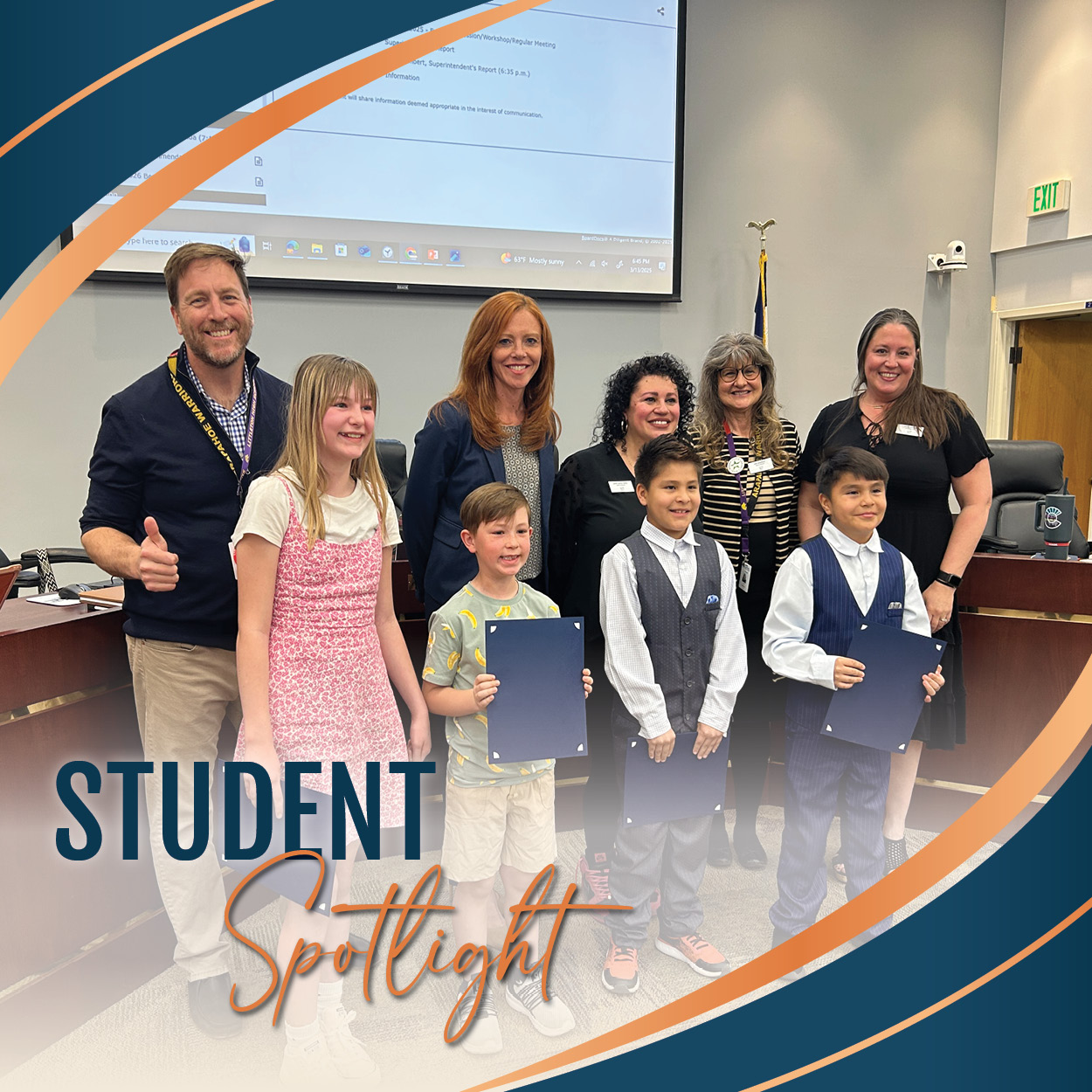 A group of 4 students stands with the Board of Education in the boardroom. The students are dressed in a mix of casual and formal attire, including two boys in matching vests and ties, a boy in a patterned shirt, and a girl in a pink floral dress. Each student is holding a certificate and smiling. The adults, dressed professionally, are standing behind them and smiling, with one man giving a thumbs-up. The background shows a projector screen and a Colorado state flag. The image includes a graphic overlay with the words "STUDENT Spotlight" in bold blue and orange text, surrounded by curved design elements in blue and orange.