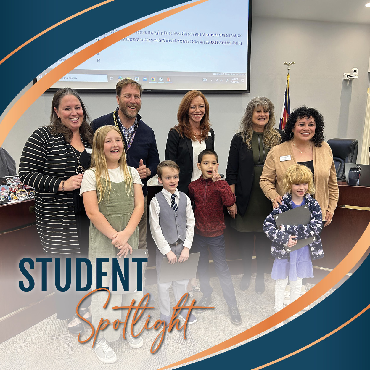 A group photo taken at a Board of Education meeting during the Student Spotlight segment. In the front row, four students, some holding certificates, smile proudly. Behind them, five Board members stand together, also smiling. A presentation screen is visible in the background, adding to the formal setting. The image is framed with a decorative border featuring blue and orange curves, adding a polished, celebratory touch.