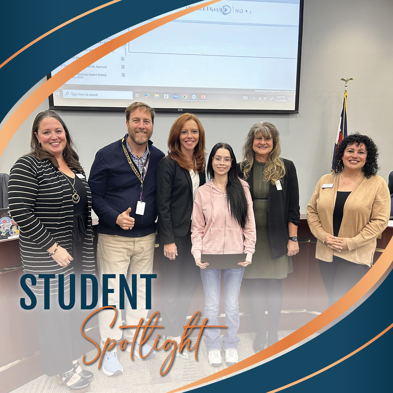 A group photo taken at a Board of Education meeting during the Student Spotlight segment. In the front row, one student holding a certificate smiles proudly. Behind the student, five Board members stand together, also smiling. A presentation screen and a flag are visible in the background, adding to the formal setting. The image is framed with a decorative border featuring blue and orange curves, adding a polished, celebratory touch.