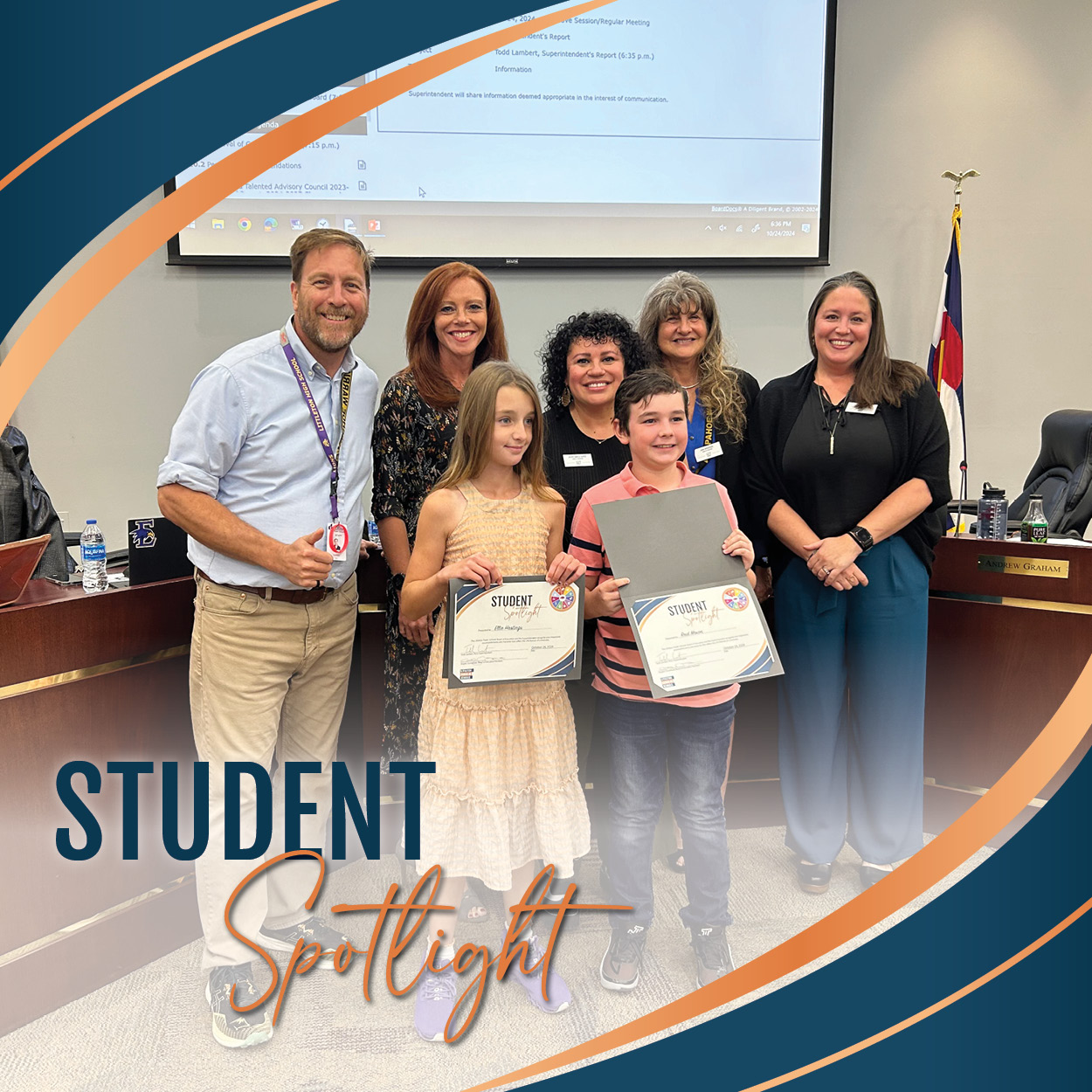 Two students holding "Student Spotlight" certificates stand proudly with members of the Board of Education at a recent board meeting, smiling for the camera. The group includes four board members standing behind the honored students. The photo is framed with a stylish blue and orange swoosh design that includes the words "Student Spotlight," adding a celebratory feel to the recognition moment.