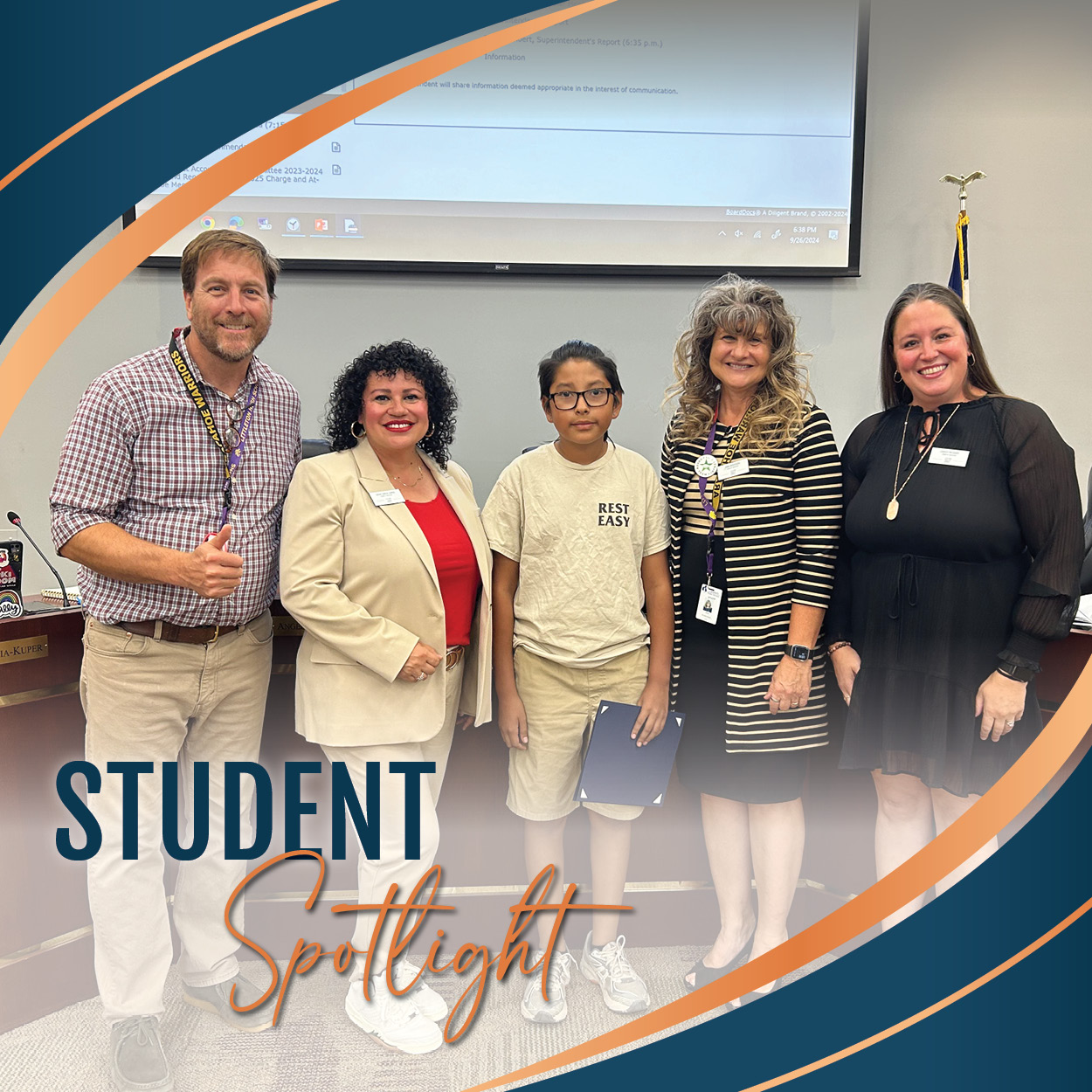 A group photo taken in a school board meeting setting. A student stands in the center, holding a certificate, dressed in a beige T-shirt and khaki shorts. Four adults, smiling, stand around the student: two women on the right and one woman and man on the left. The man gives a thumbs up, and everyone appears to be celebrating the student's recognition. The text 'Student Spotlight' is displayed in a bold and elegant font at the bottom left of the image with a colorful, curved graphic design framing the photo.