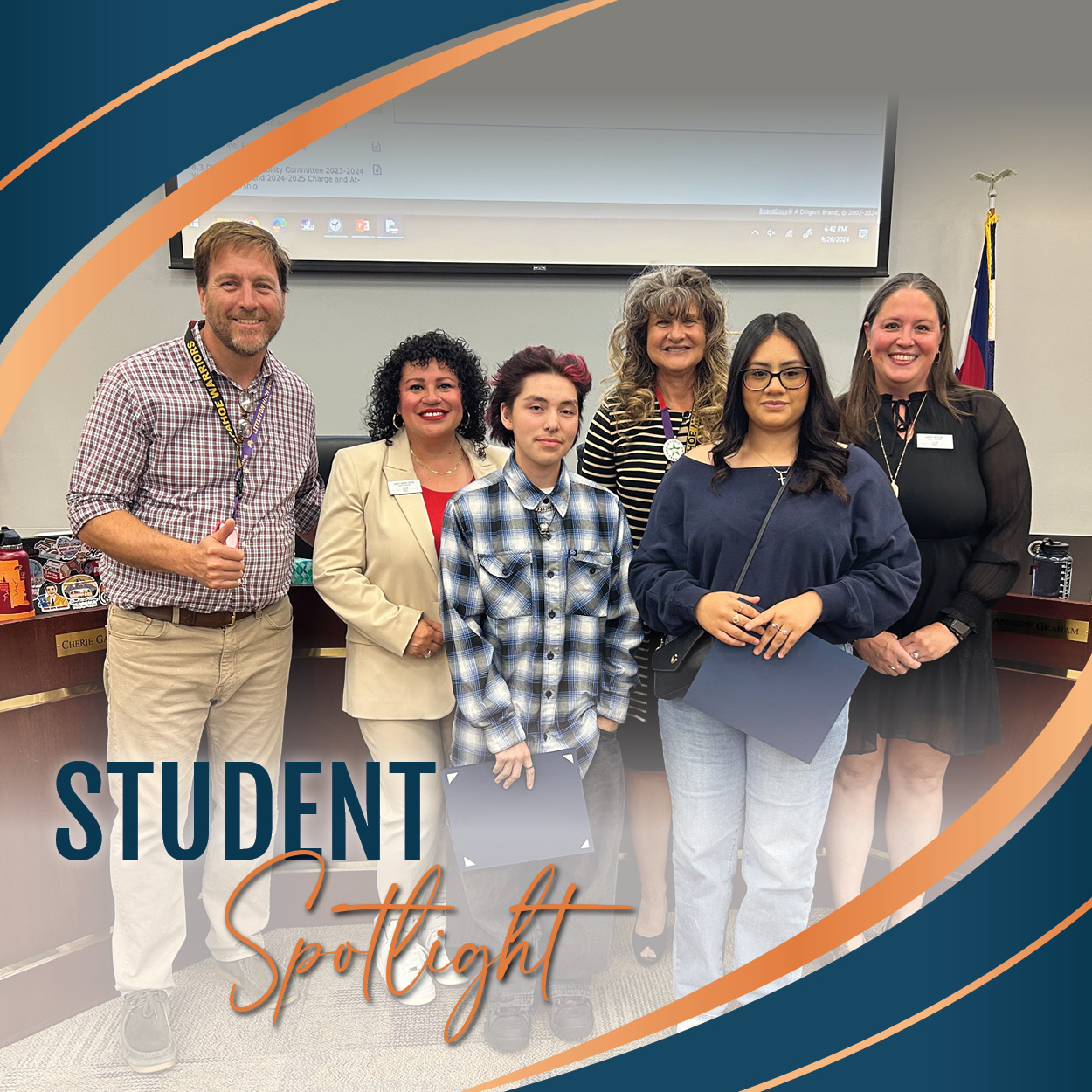 A group photo taken in a school board meeting setting. Two students stand in the center, both holding certificates. One student is wearing a blue plaid shirt and the other a blue sweatshirt with glasses. They are surrounded by four smiling adults: two women on the right and one woman and man on the left. The man on the far left gives a thumbs up. The text 'Student Spotlight' is displayed in a bold and elegant font at the bottom left of the image with a colorful, curved graphic design framing the photo.