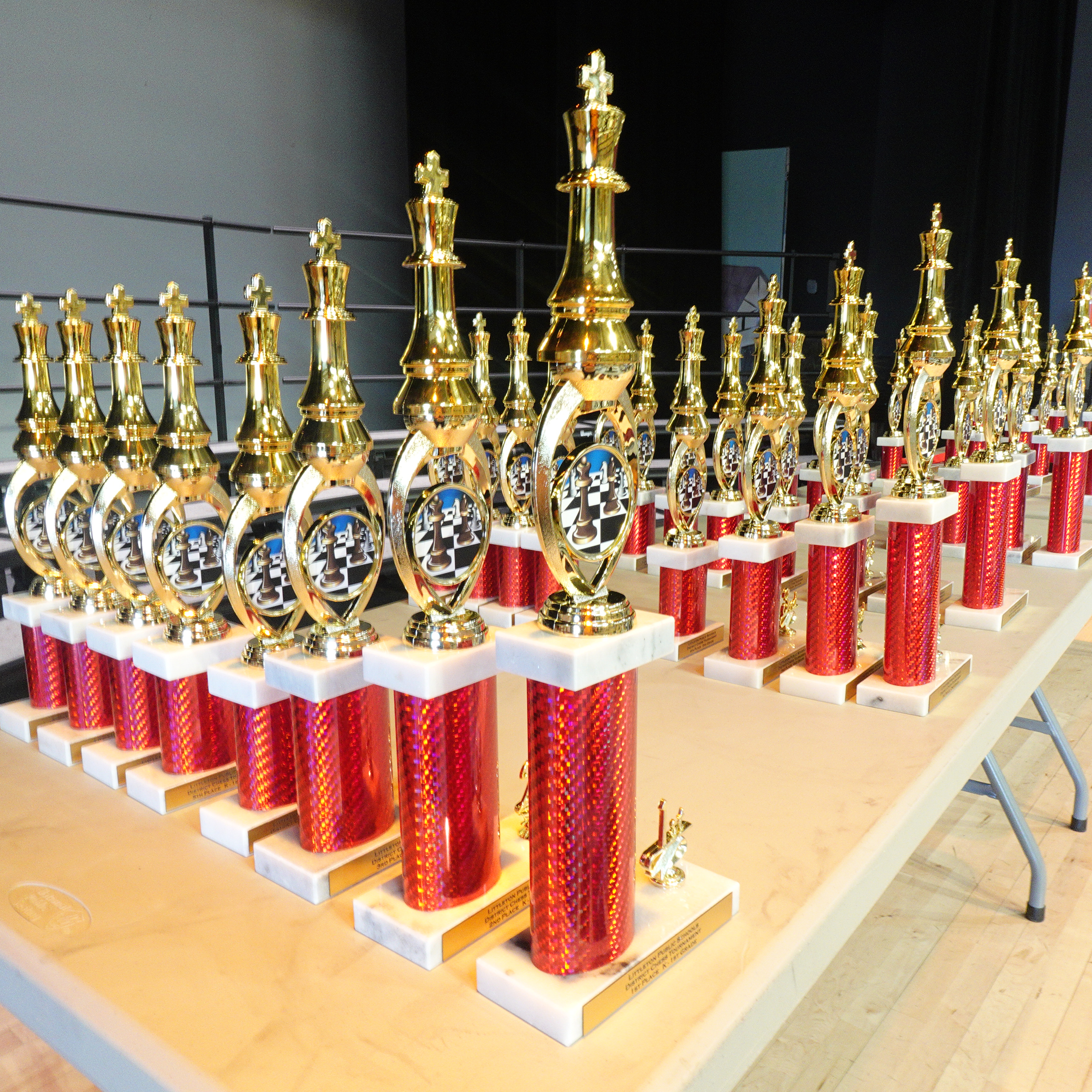 Rows of gleaming red and gold chess trophies displayed on a table, each featuring a chess piece design on top, ready to be awarded to participants.